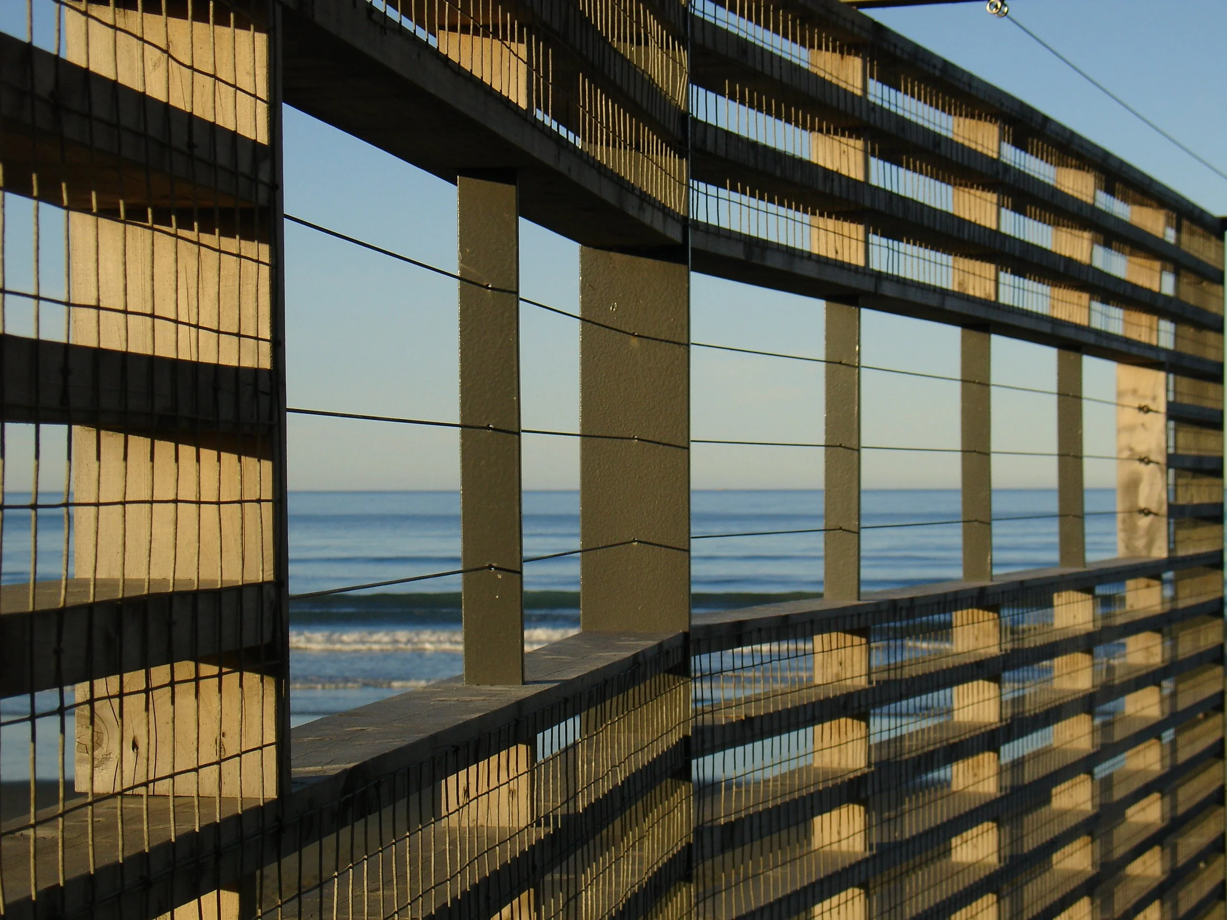 View of the ocean through a wooden and wire fence on a pier or boardwalk at sunset.