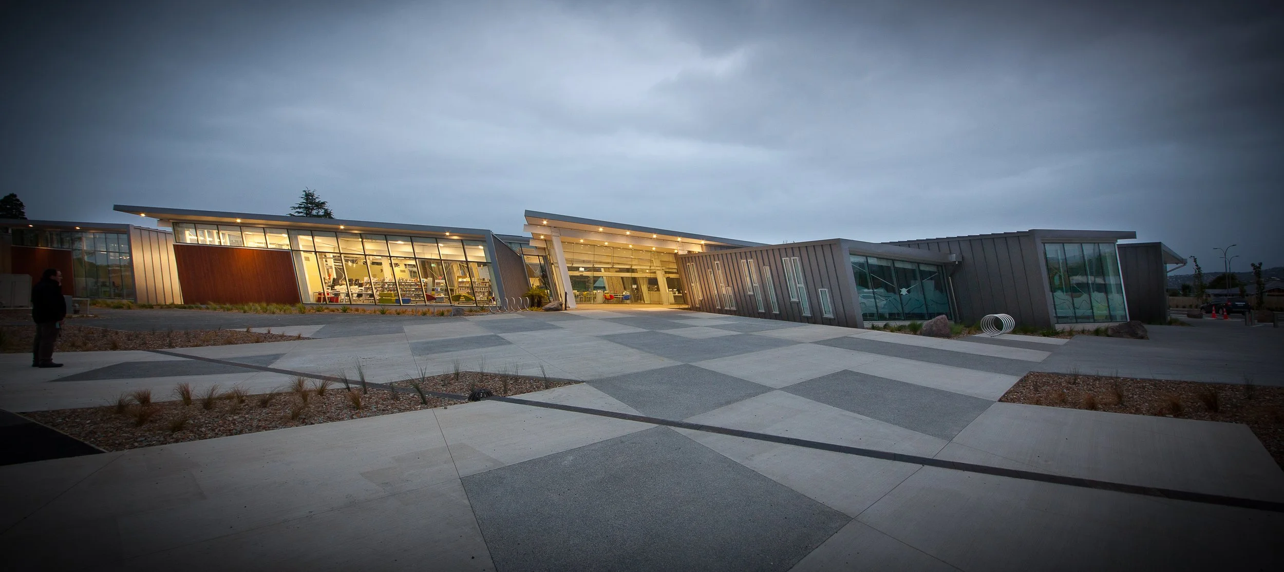 Modern building with large glass windows, illuminated from inside, under a cloudy evening sky. The architecture features angular designs and a patterned concrete pavement in the foreground.