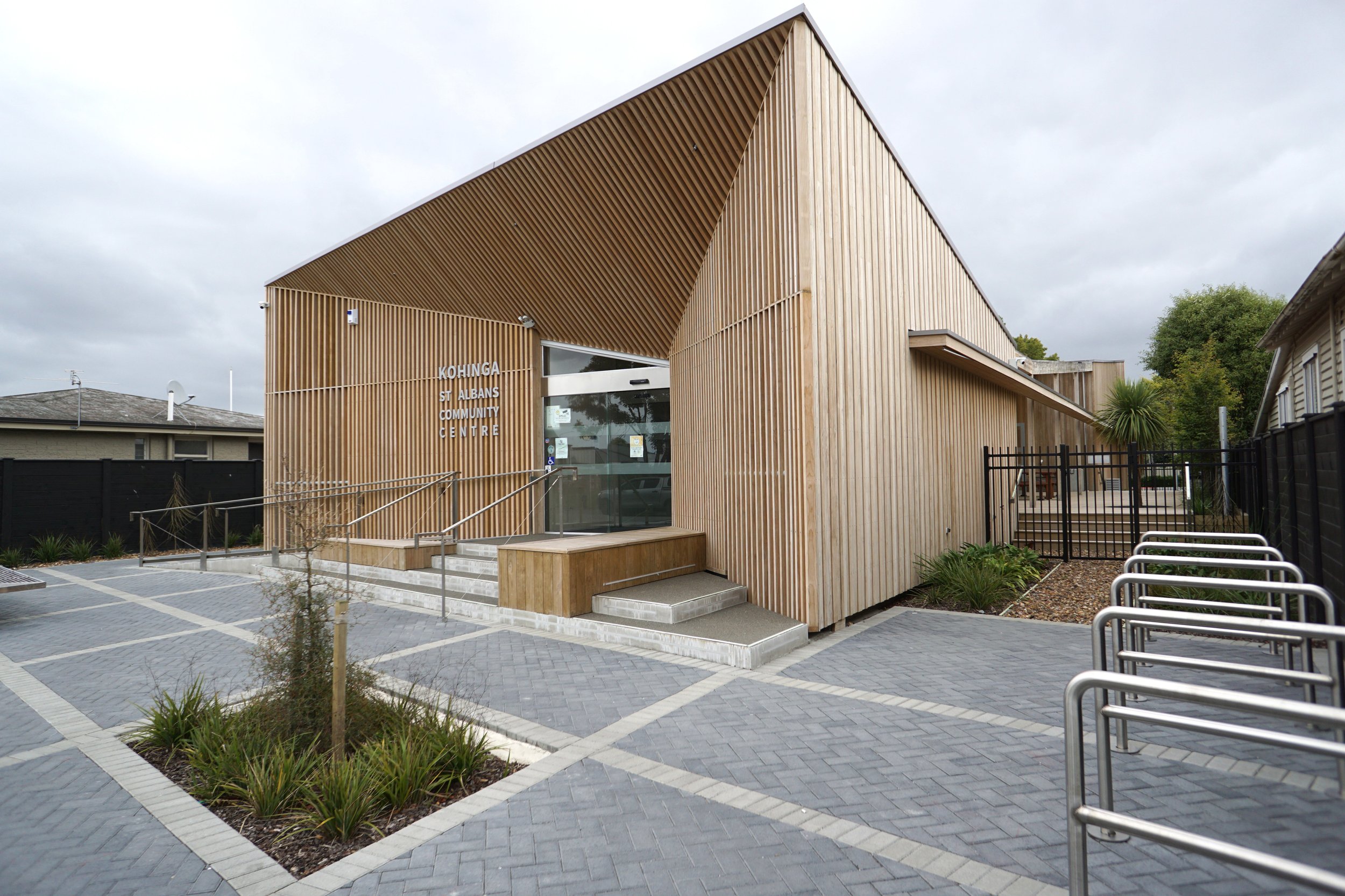Modern wooden building labeled Kohinga St Albans Community Centre with wide steps, designated disabled parking, and bike racks in front, surrounded by a paved area and small landscaped garden beds.