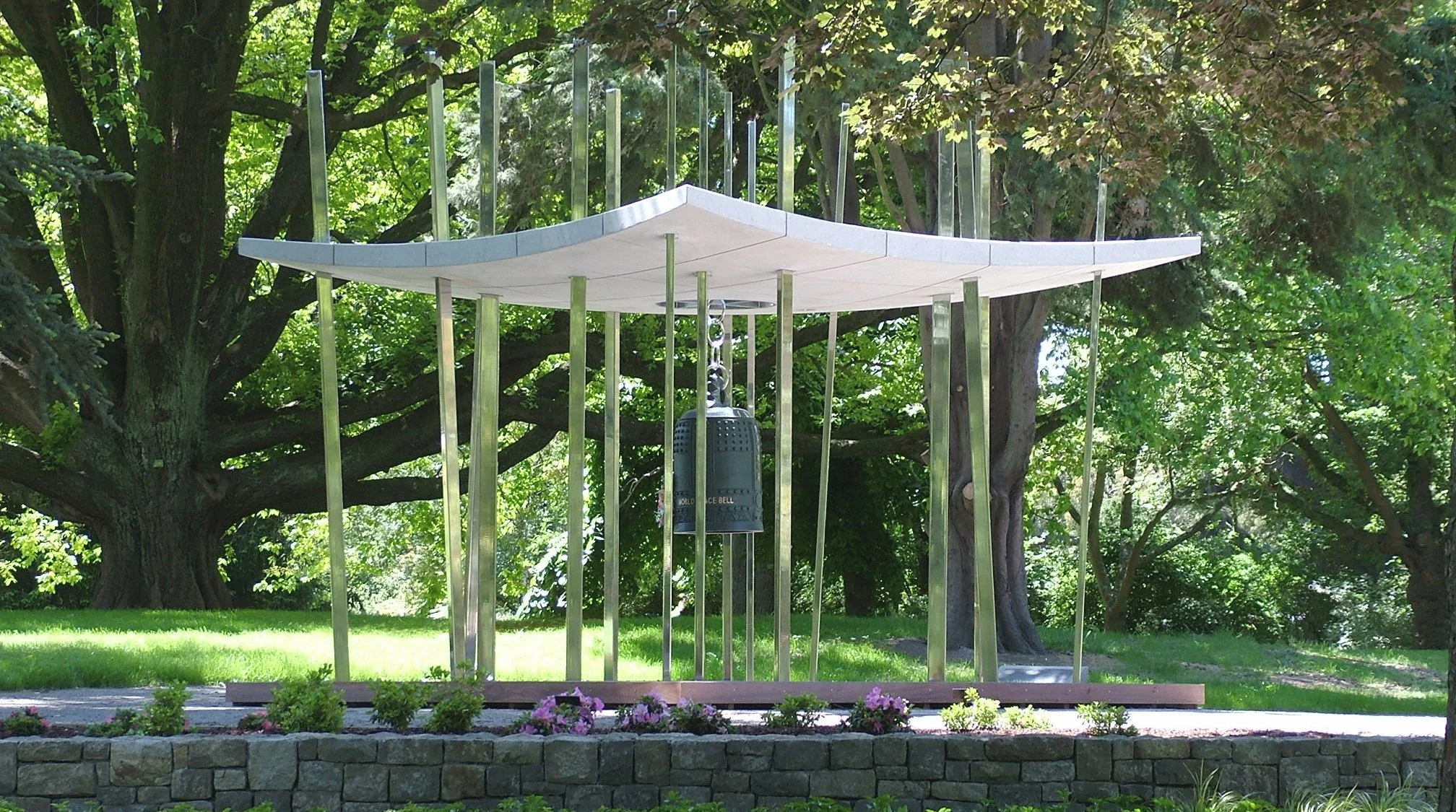 A modern monument with a curved, white roof supported by thin metal supports, with a bell hanging in the center, situated in a park with green trees and flowers.