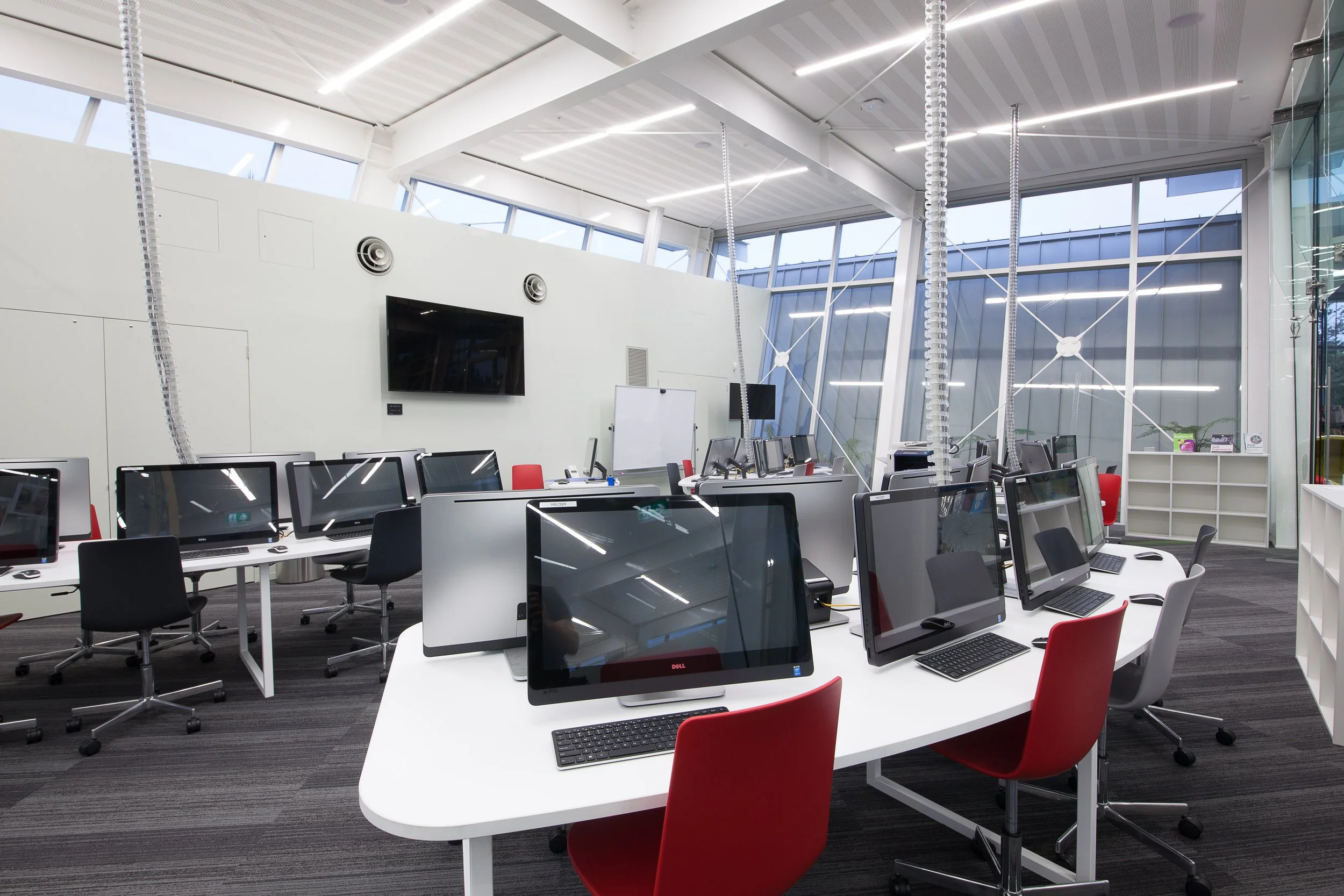 Modern office with multiple computer stations, white desks, black and red chairs, large windows, wall-mounted TV, whiteboard, and ceiling lighting.