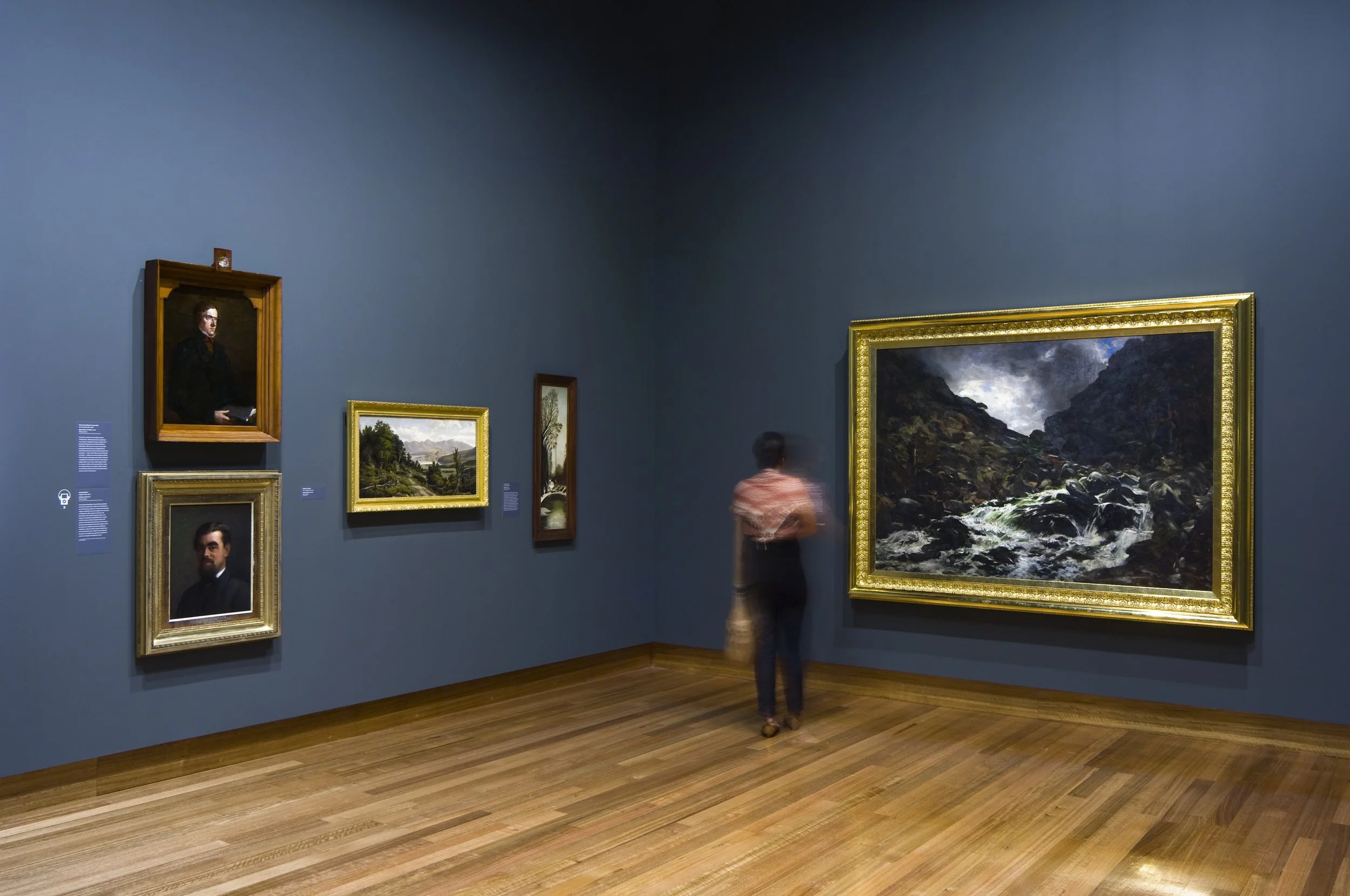 A person finds and looks at landscape paintings in an art gallery with dark blue walls and wooden floors.