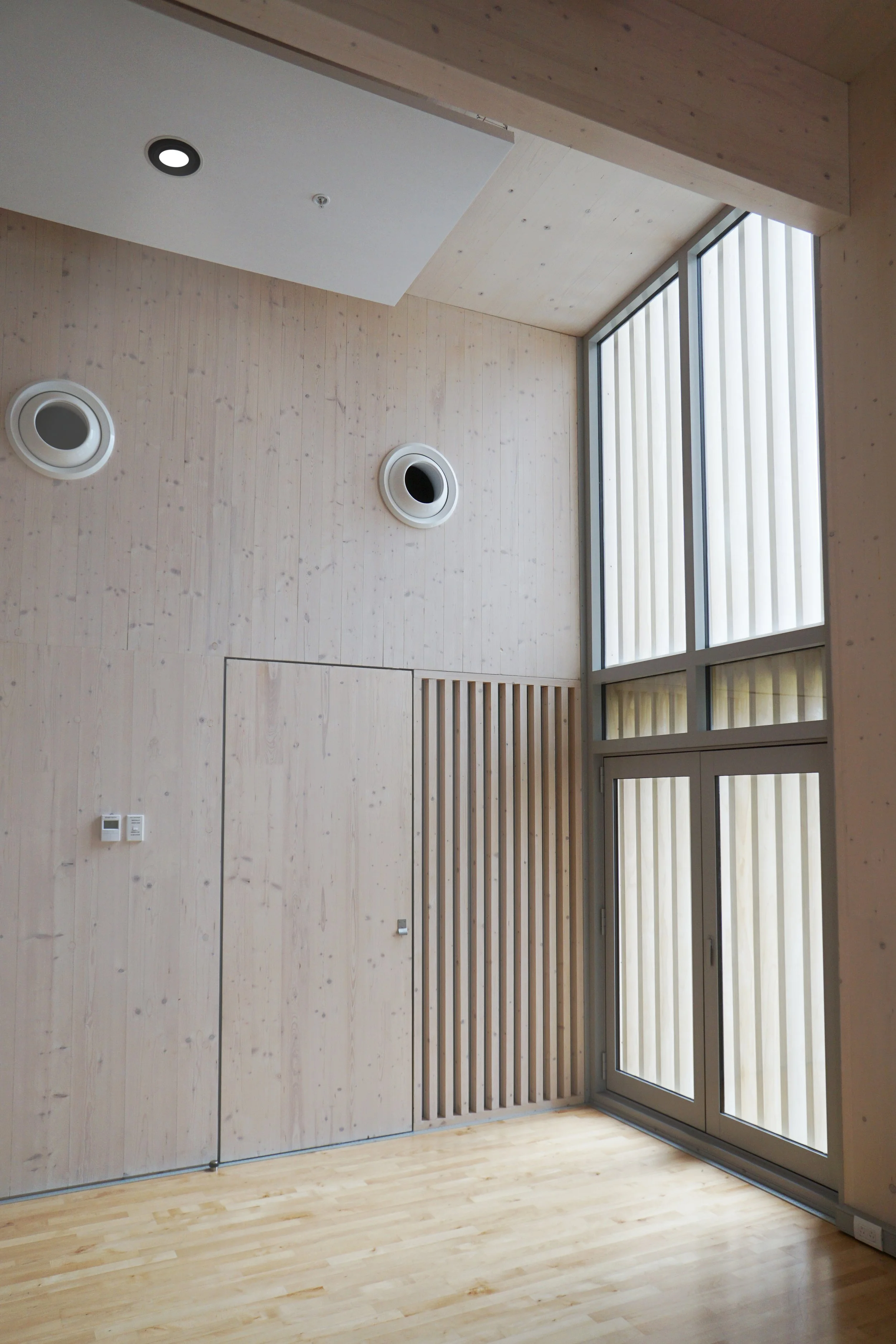 Interior view of a modern building with light wood panel walls, two circular ventilation openings in the upper wall, large glass windows with vertical blinds, wooden flooring, and a door integrated into the wall.