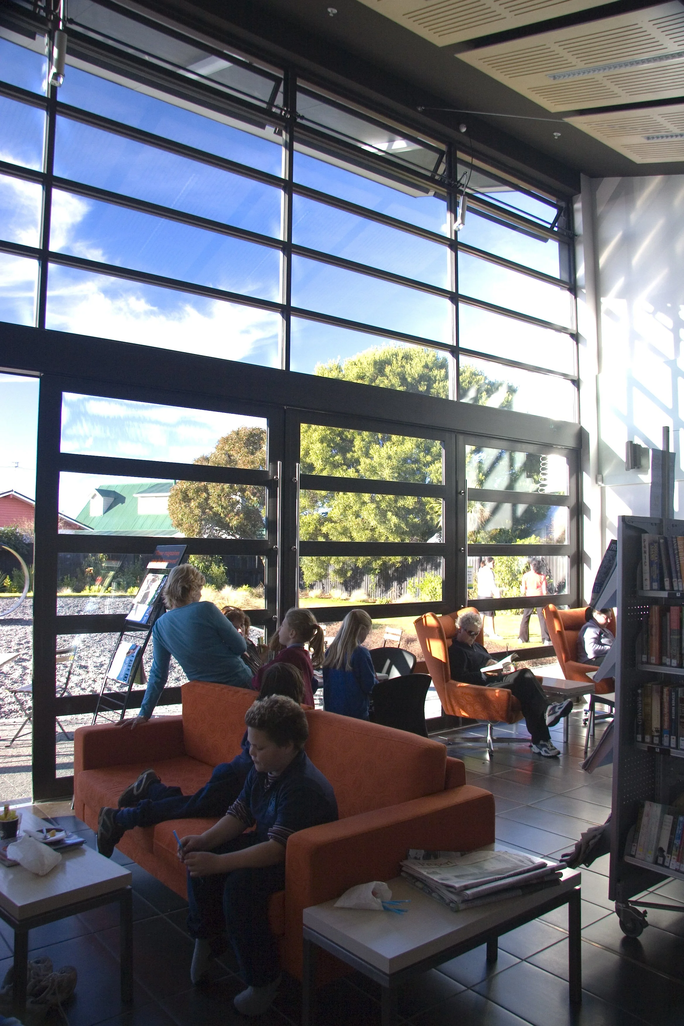 Interior of a library with large windows, several people sitting in chairs and on a sofa, reading or using devices, with sunlight streaming in.