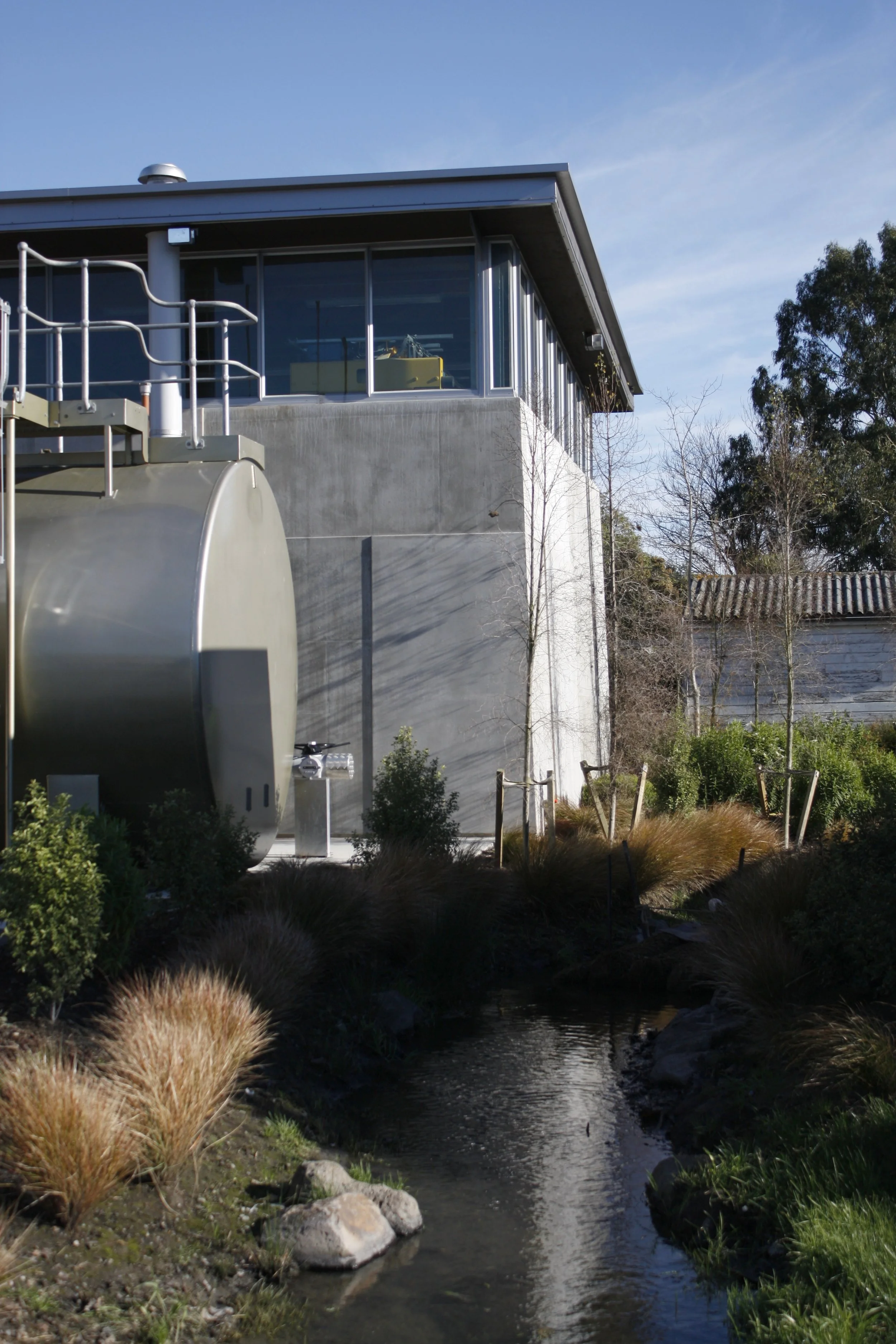 Modern building with large windows, a cylindrical metallic tank, and a small stream of water surrounded by plants and trees.