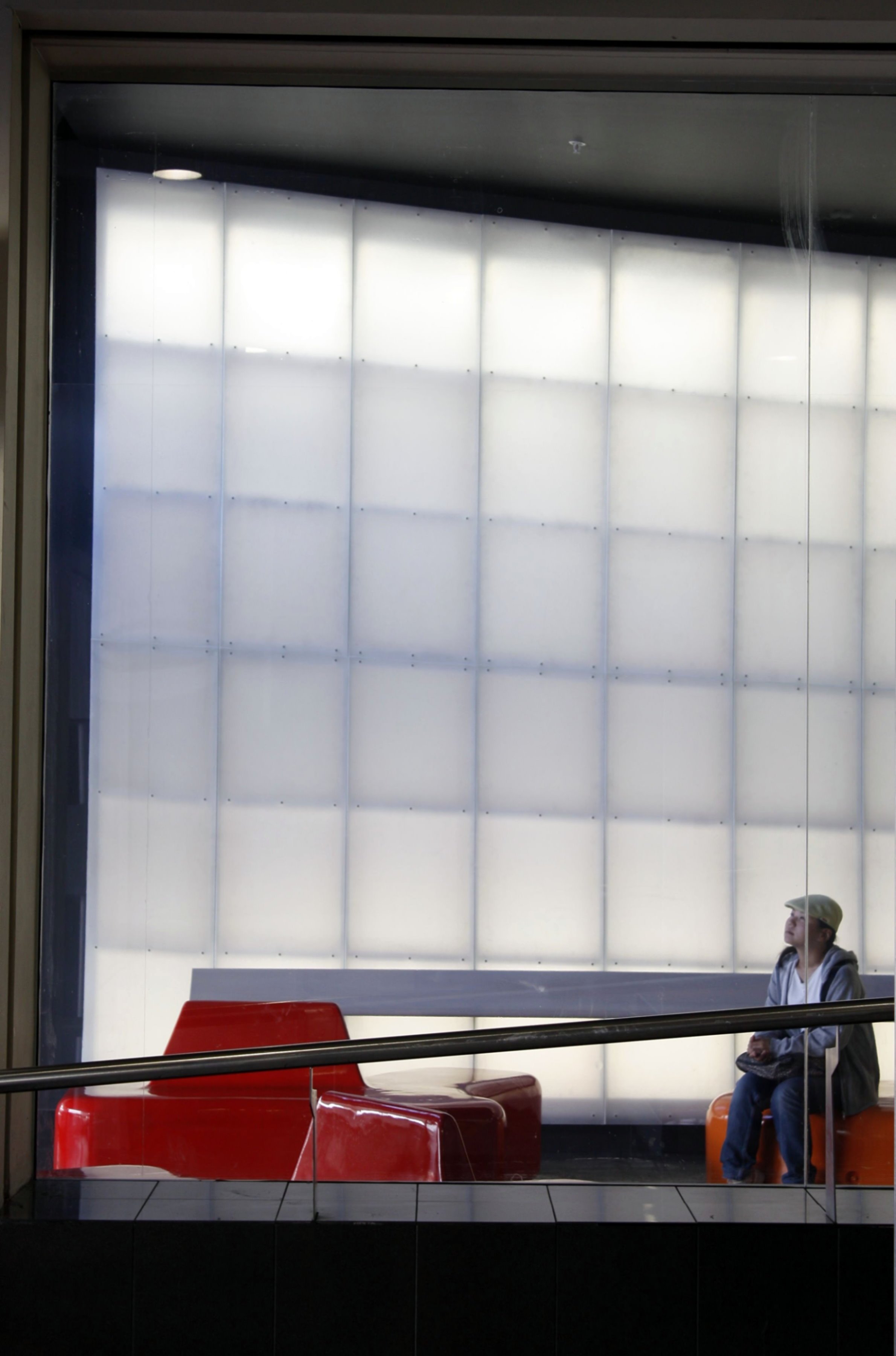 A person sitting on an orange bench, holding a phone, near red chairs, viewed through a glass window with a frosted white background.