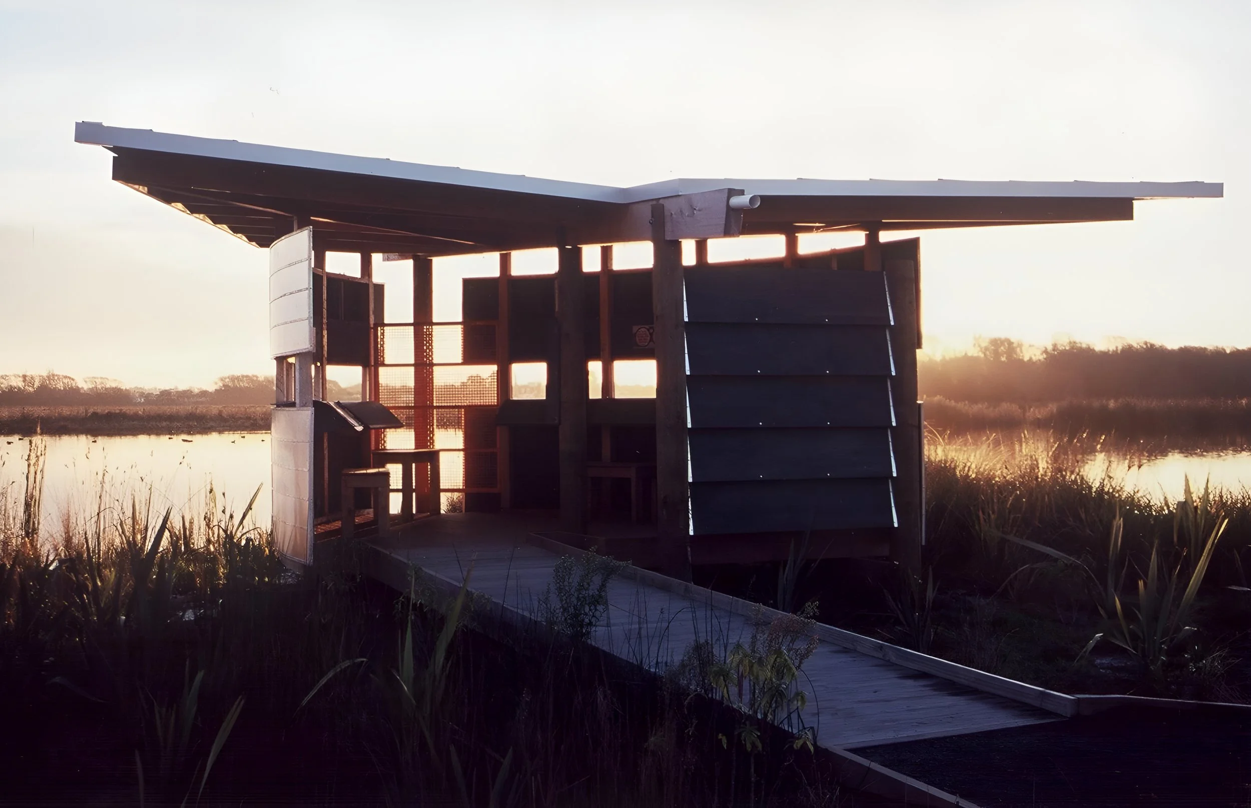 A wooden shelter with an overhanging roof on a boardwalk, located near a marsh or wetland at sunset.