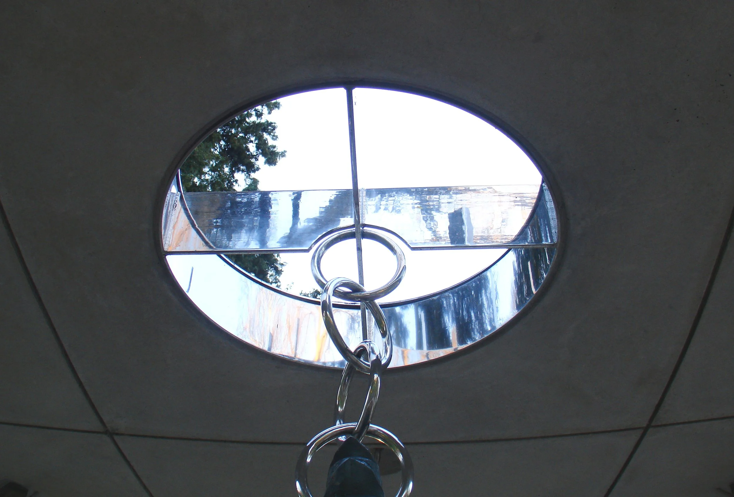 Looking up through a circular skylight with a metal ring handle in the center, and the sky and trees visible outside.