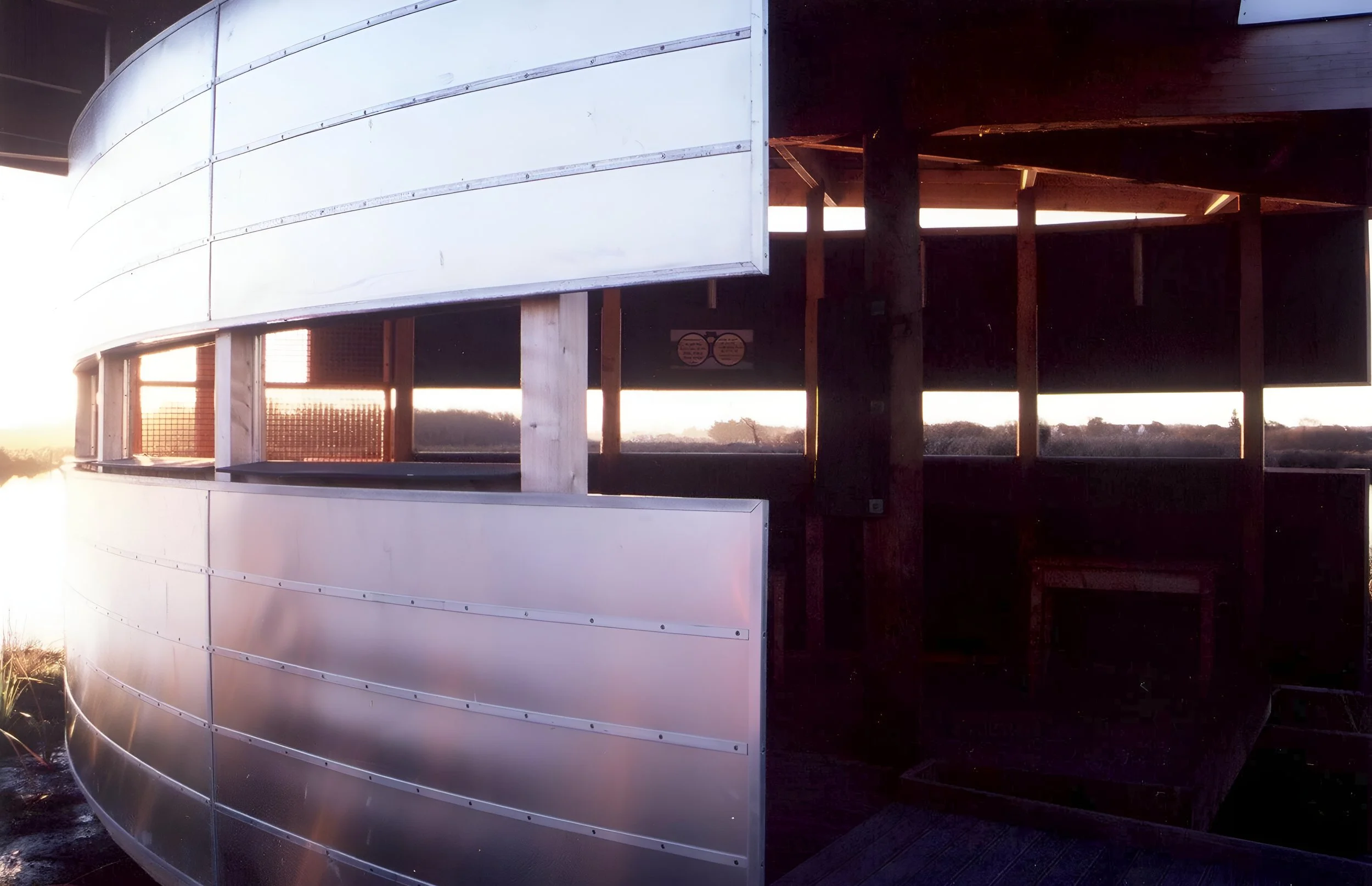 Partially constructed modern building with curved, metallic exterior panels and open windows, with a sunset in the background.