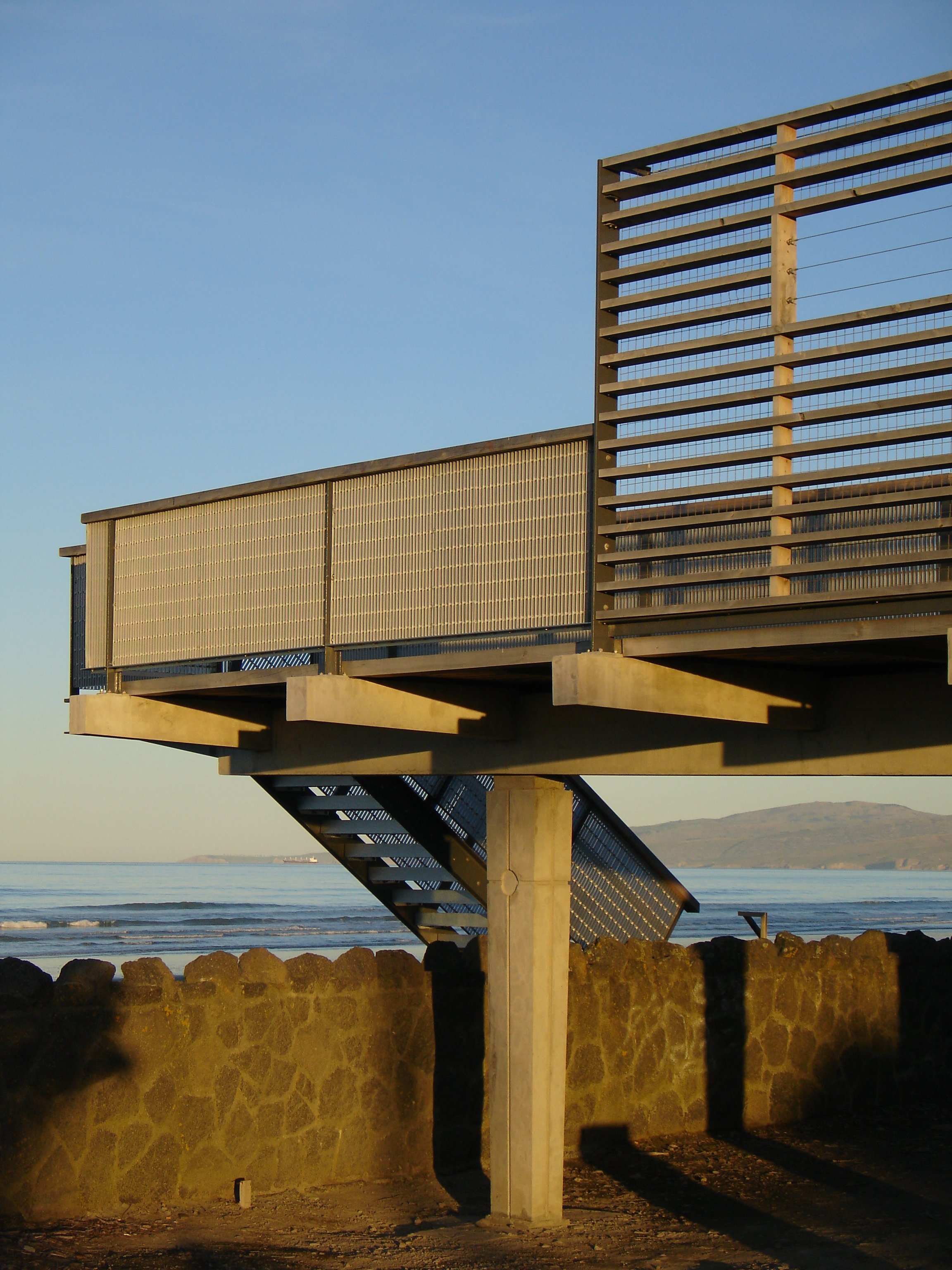 Beachside modern structure with metal stairs and orange-tinted wood slats, overlooking ocean, with distant hills in the background.