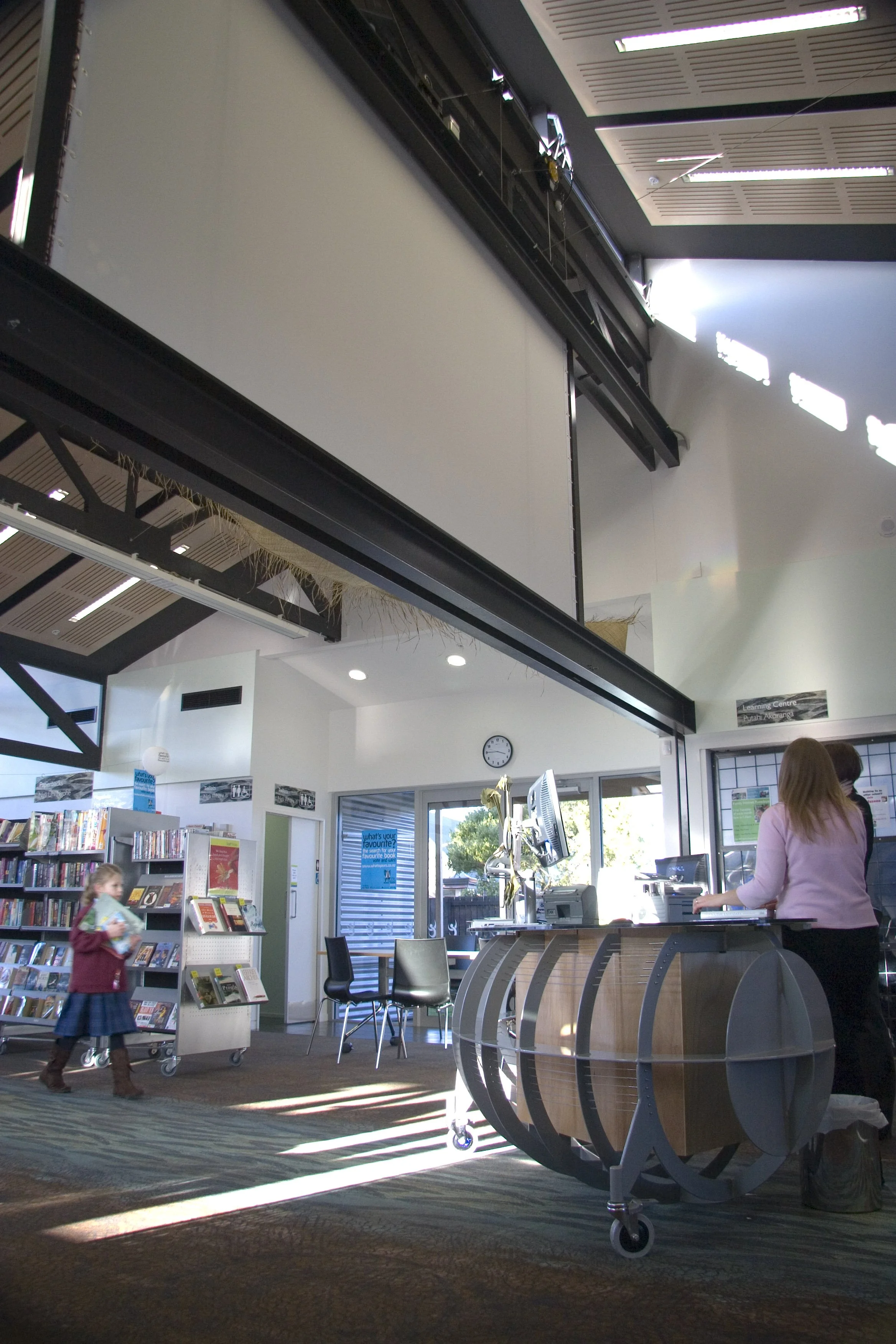 Interior of a library with a reception desk, a child holding books walking, and a woman working at the counter. There are bookshelves, chairs, and large windows letting in natural light.