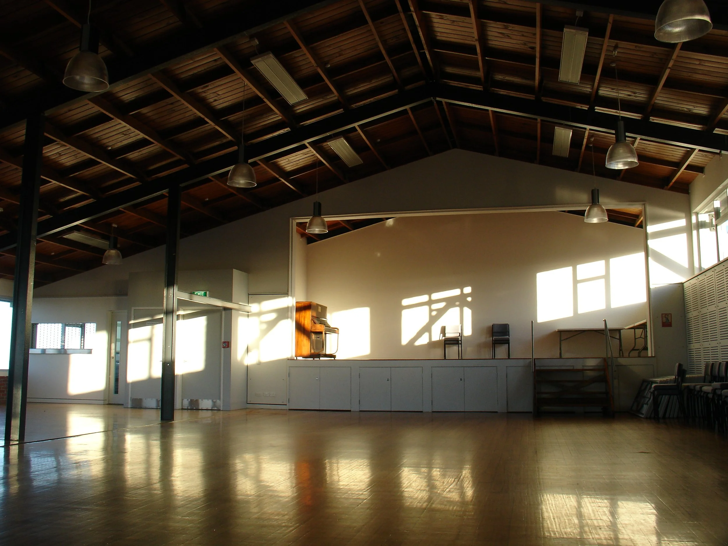 An empty spacious hall with a high wooden ceiling, sunlight streaming through windows, a small stage with two chairs, a piano, and stacked chairs on the side.