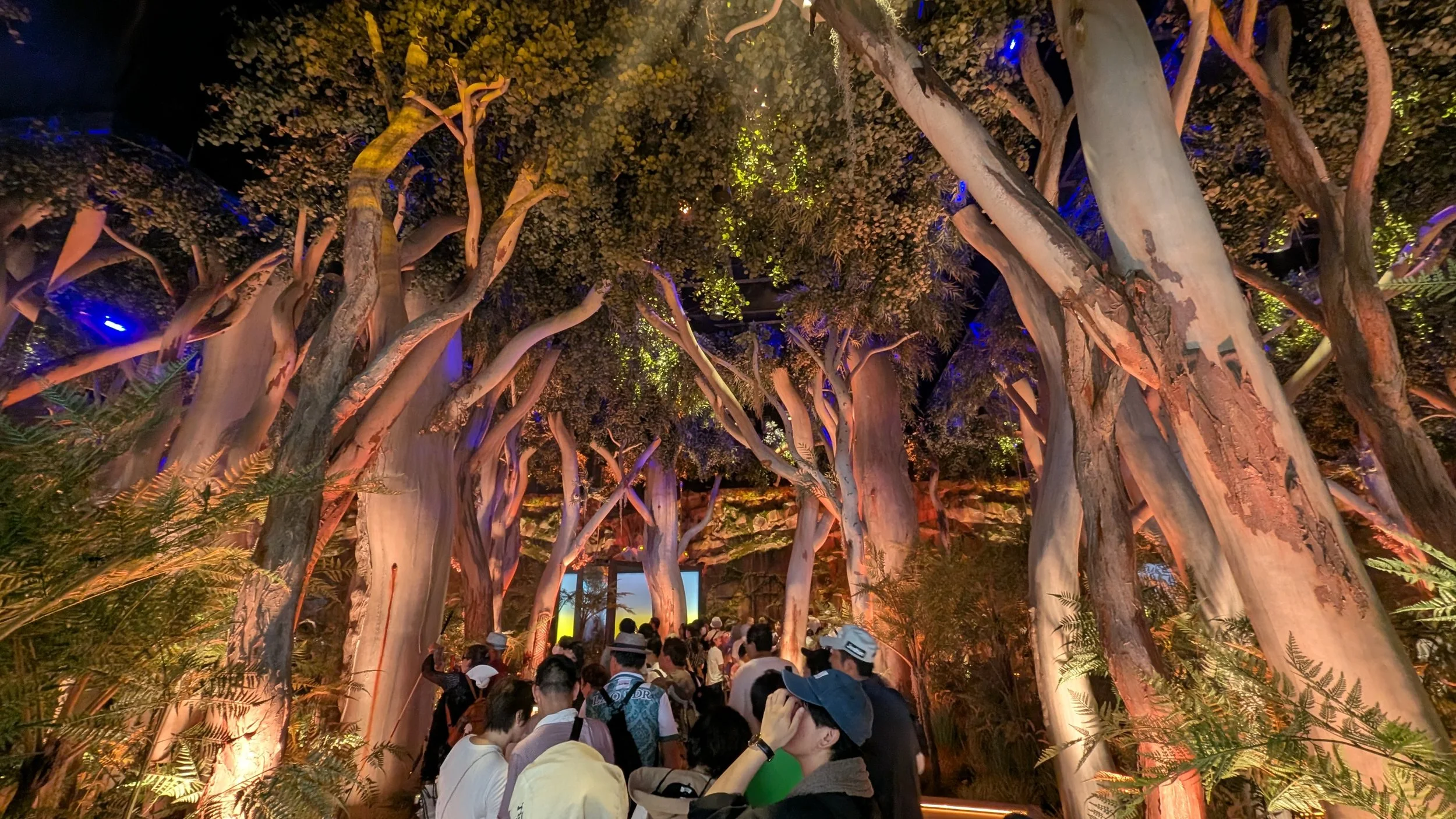 People gathered inside a large indoor space with tall, twisted artificial trees and foliage, under colorful lighting.