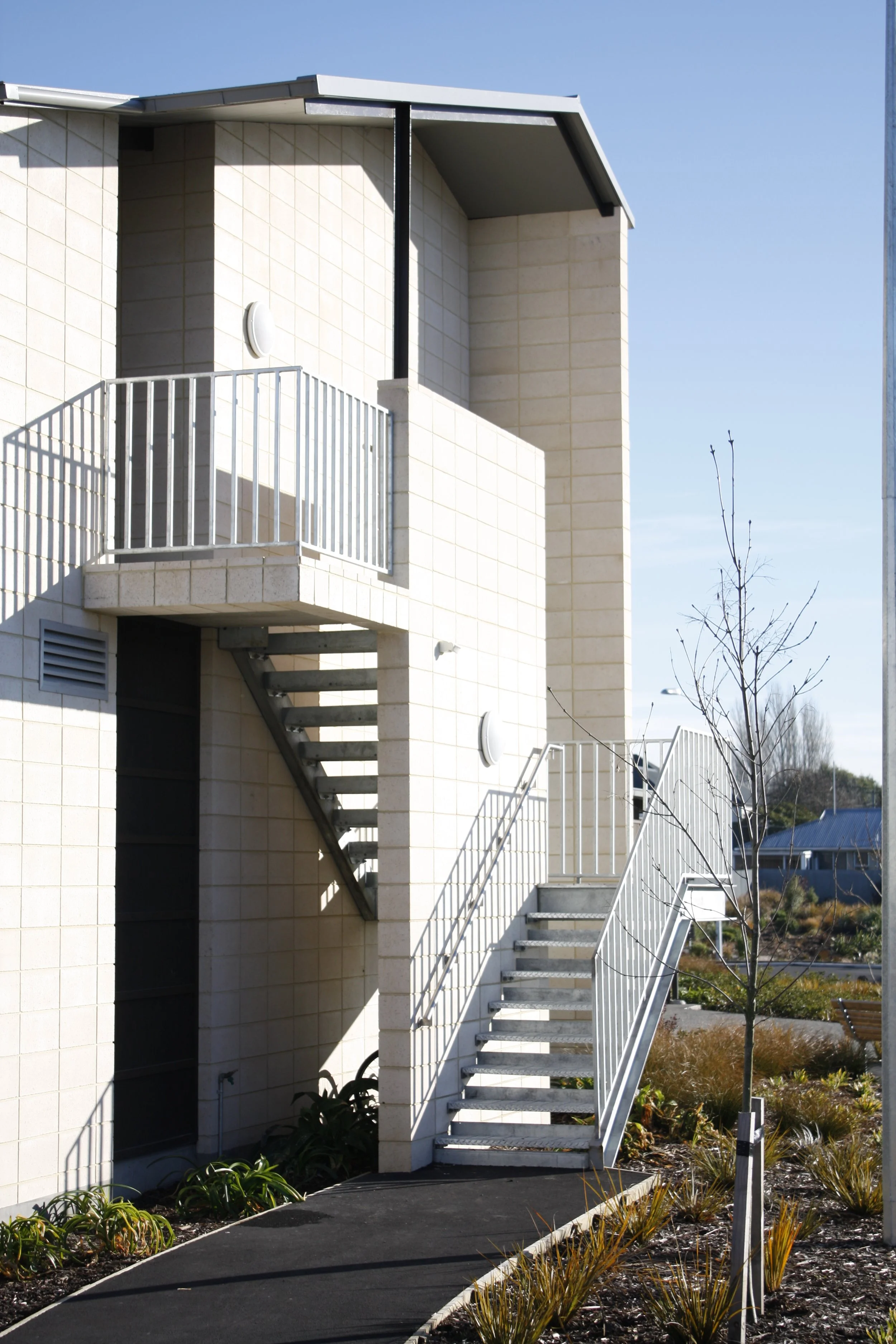 Exterior view of a modern building with metal staircase, small balcony, and minimal landscaping, under a clear blue sky.