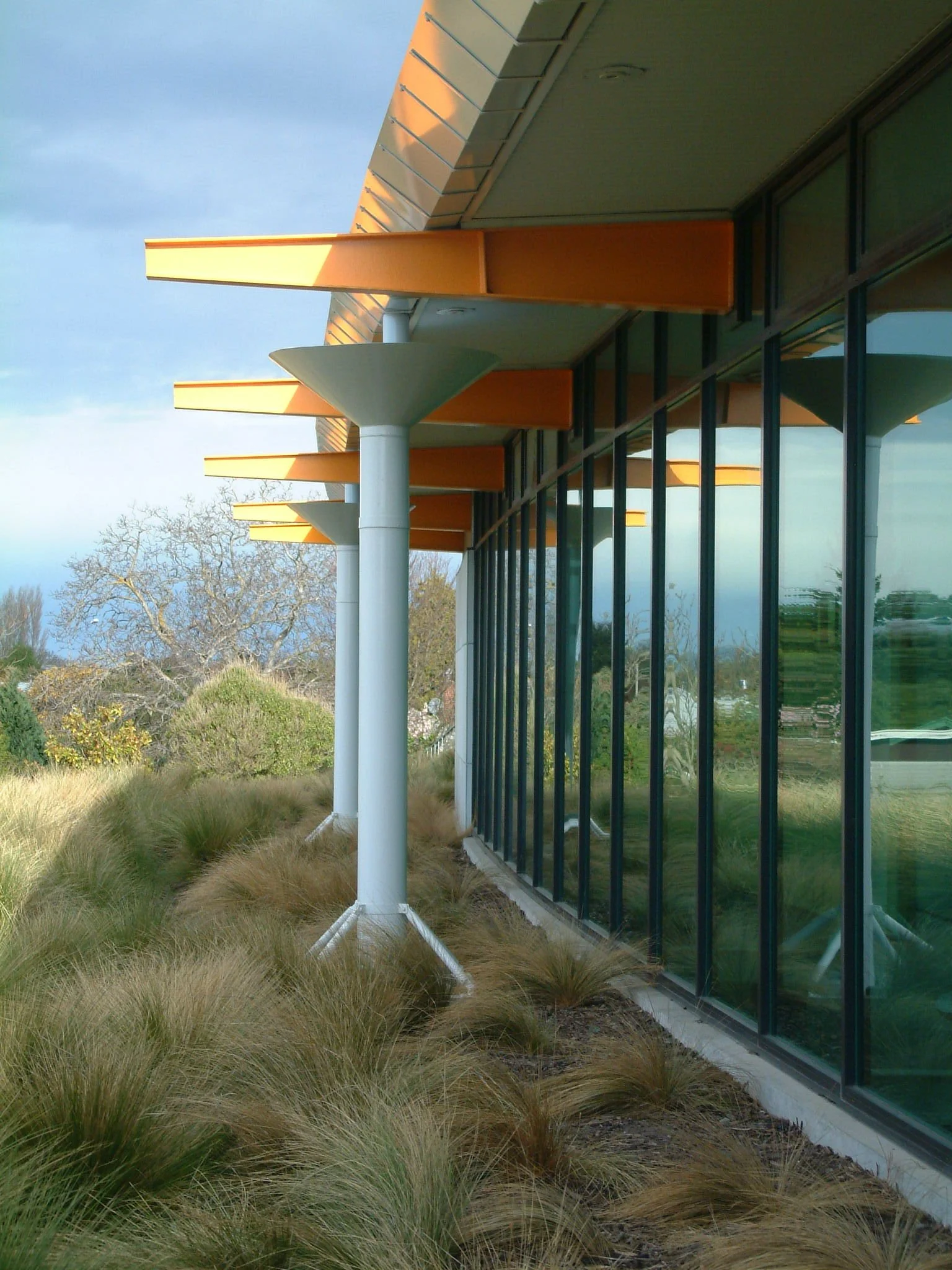 Exterior view of a modern building with large glass windows, orange architectural canopies, and white supporting columns, with ornamental grasses in the landscaping.