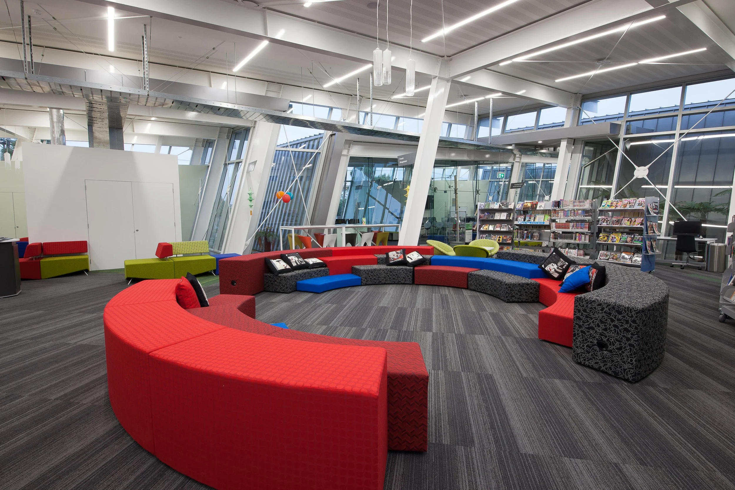 Modern library or lounge area with colorful curved seating, bookshelves, large windows, and contemporary lighting fixtures.