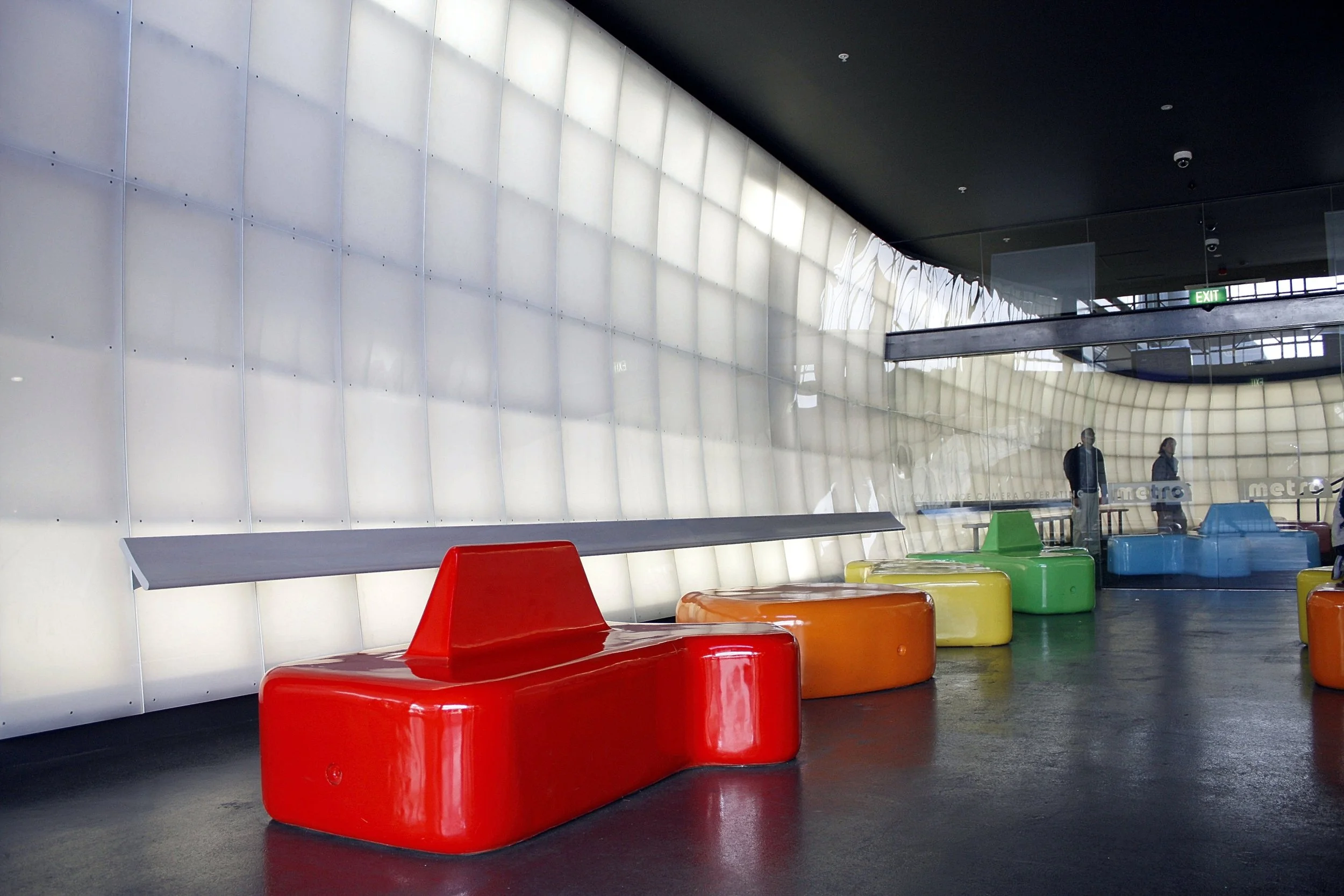 Colorful modern benches in front of a large illuminated wall and glass window at a public transportation station, with two people walking in the background.