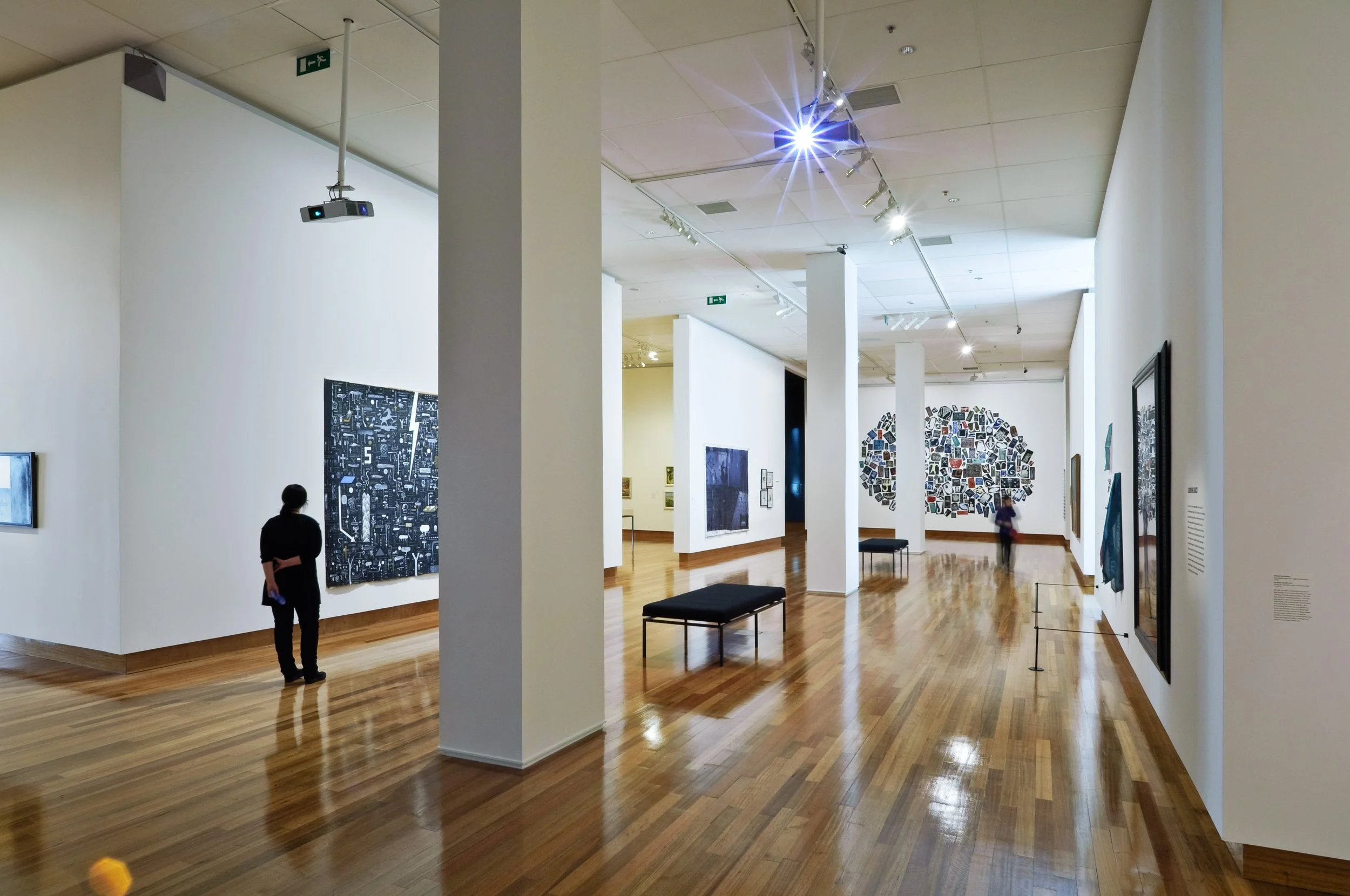 Interior of an art gallery with wooden flooring, white walls displaying various artworks, and a few visitors observing the art pieces.