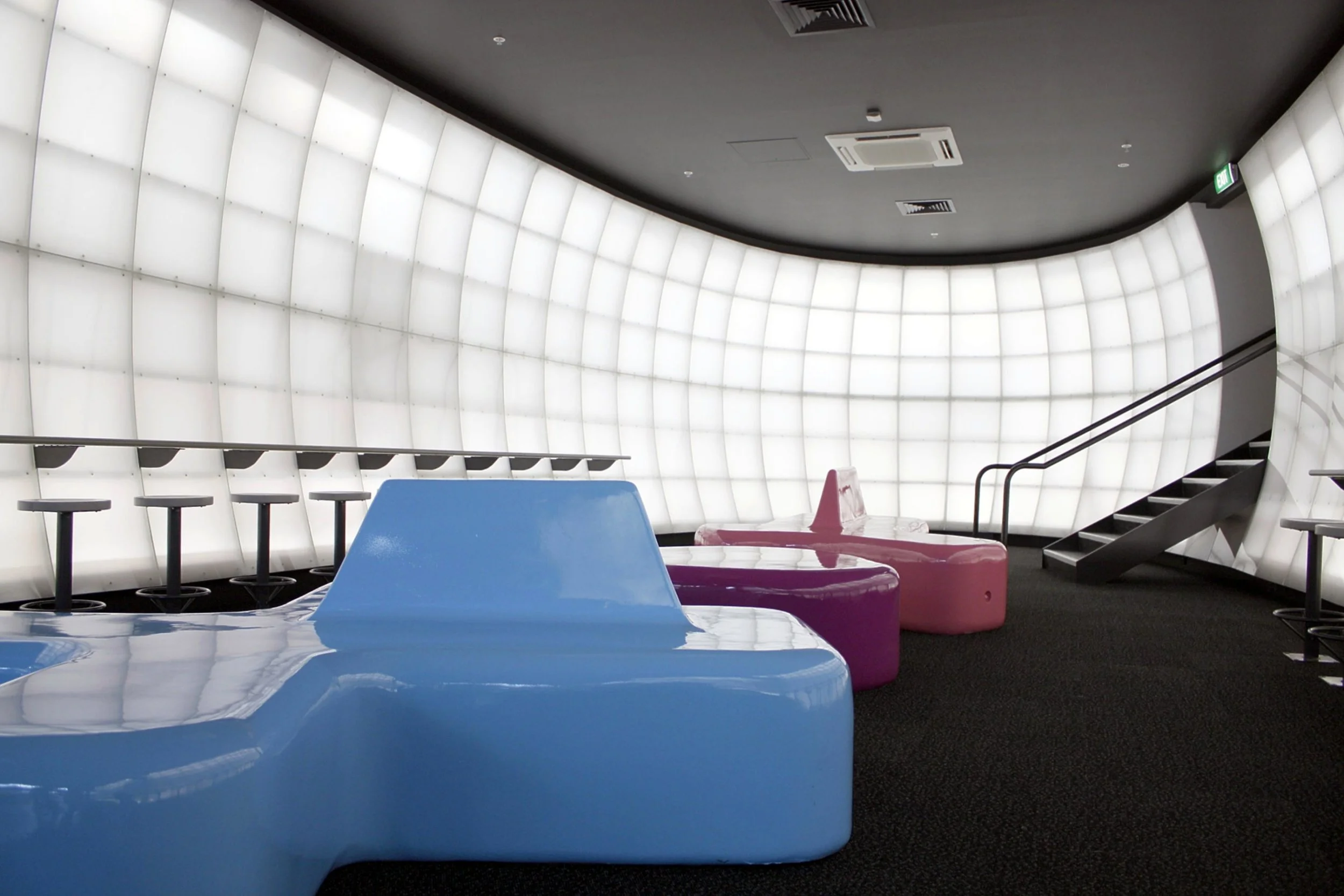 Modern interior space with a curved illuminated white wall, black floor, and colorful plastic chairs in blue and pink, along with barstools against the wall and a staircase leading up on the right side.