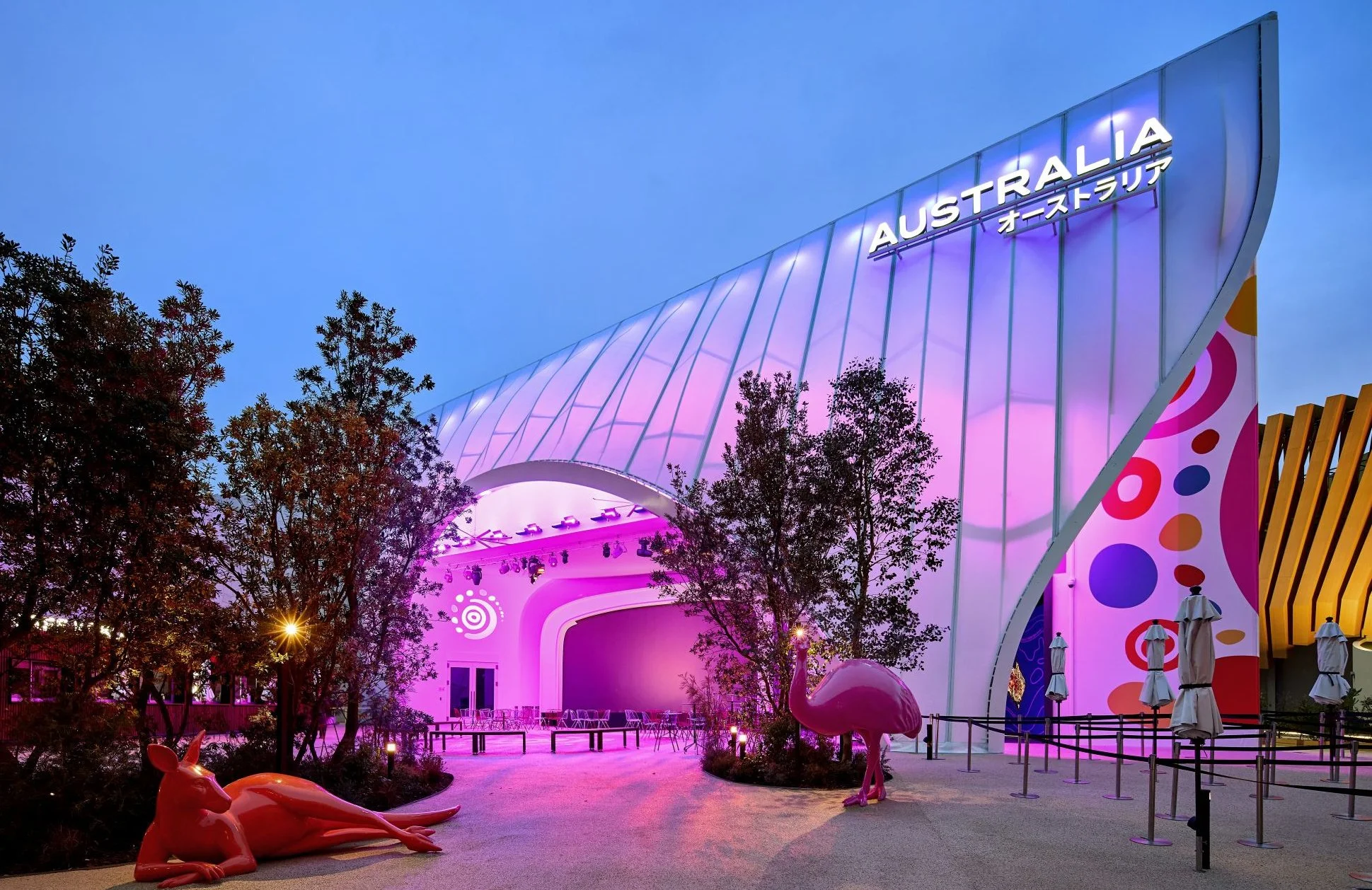 The image depicts a modern, colorful building under a blue sky with the word "Australia" illuminated on the top. There are decorative pink flamingo and animal sculptures in the foreground, with trees and umbrellas nearby. The building has a curved, f