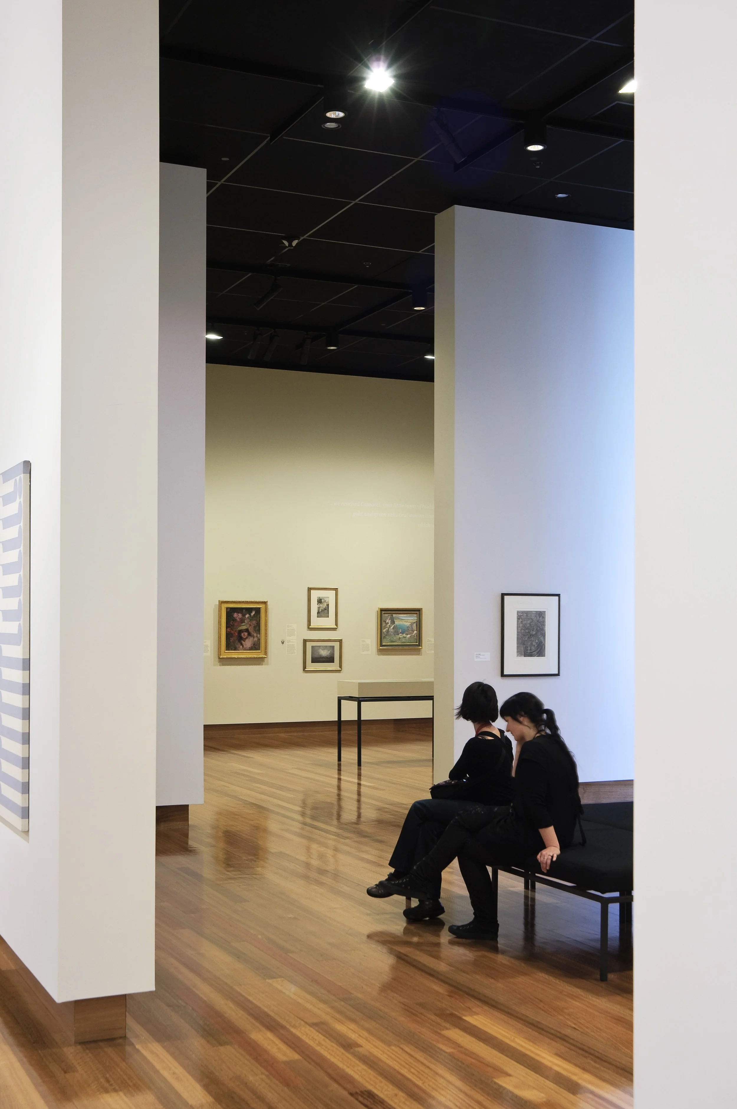 Two women sitting on a bench in an art gallery, with framed artwork on the wall in the background.