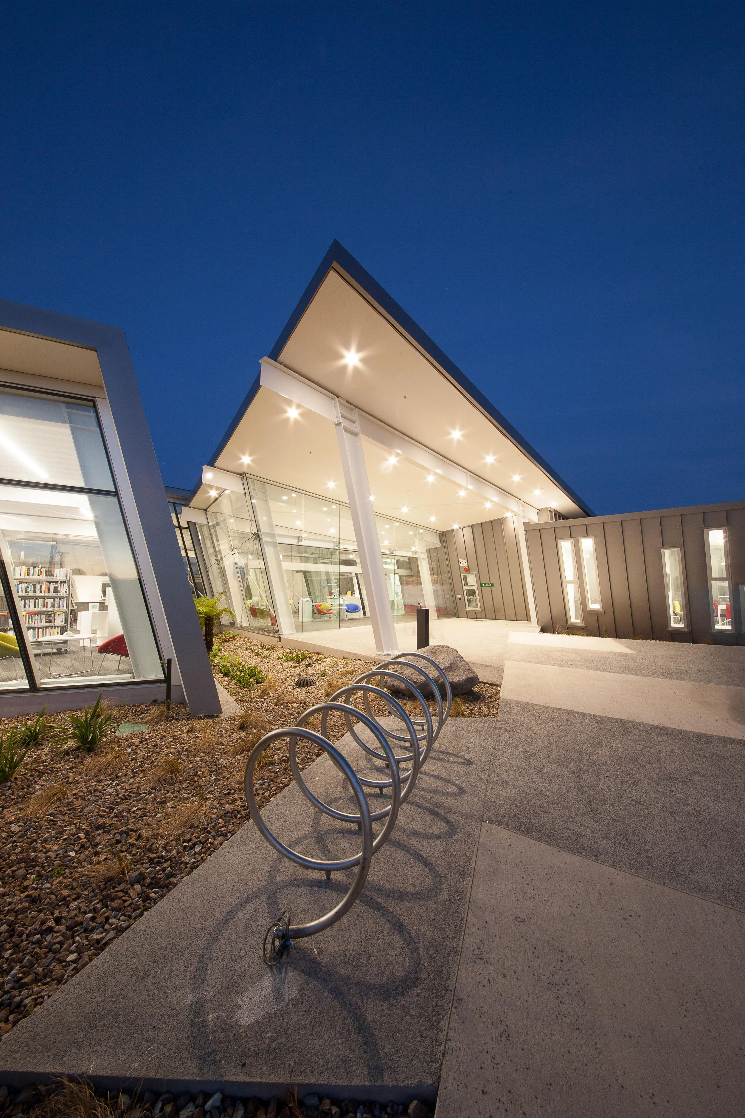 Modern architectural building with large glass windows and a sloped roof, illuminated at night, with bike racks in the foreground.