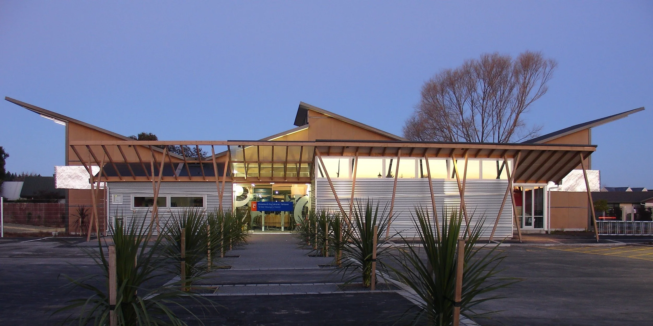 Modern building with angular roof design, illuminated from inside, surrounded by parking lot and landscaped plants, with leafless trees in background at dusk.