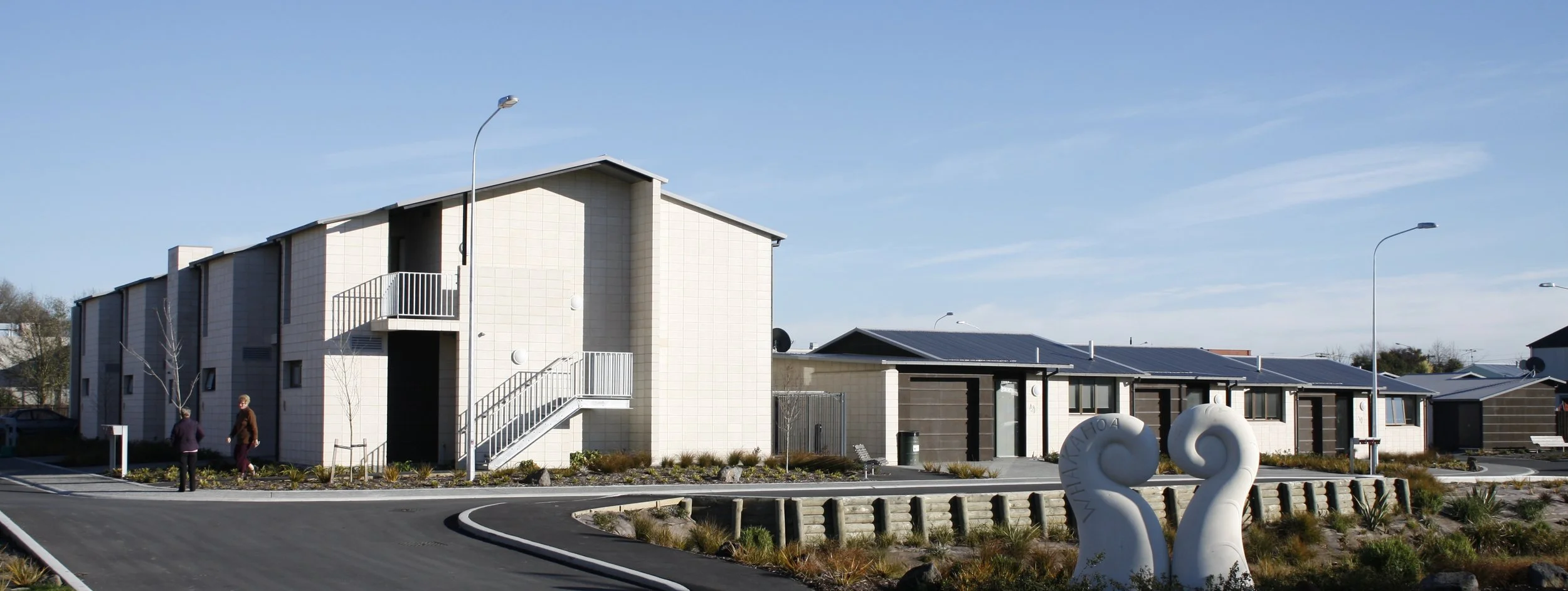 A modern residential area with multiple buildings, some featuring solar panels on the roofs. Two people are walking on a curved paved pathway, with landscaped plants and decorative sculptures in the foreground. The sky is clear and blue.