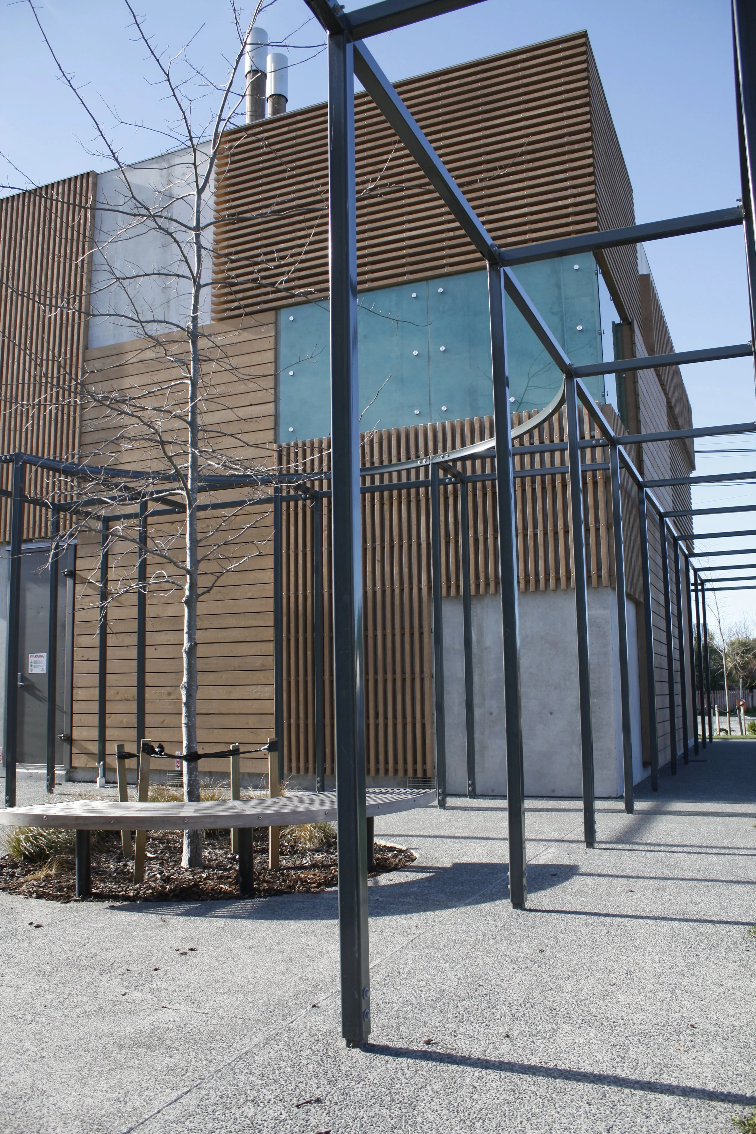Modern building exterior with wooden and concrete panel design, metal fencing, leafless trees, and clear blue sky.