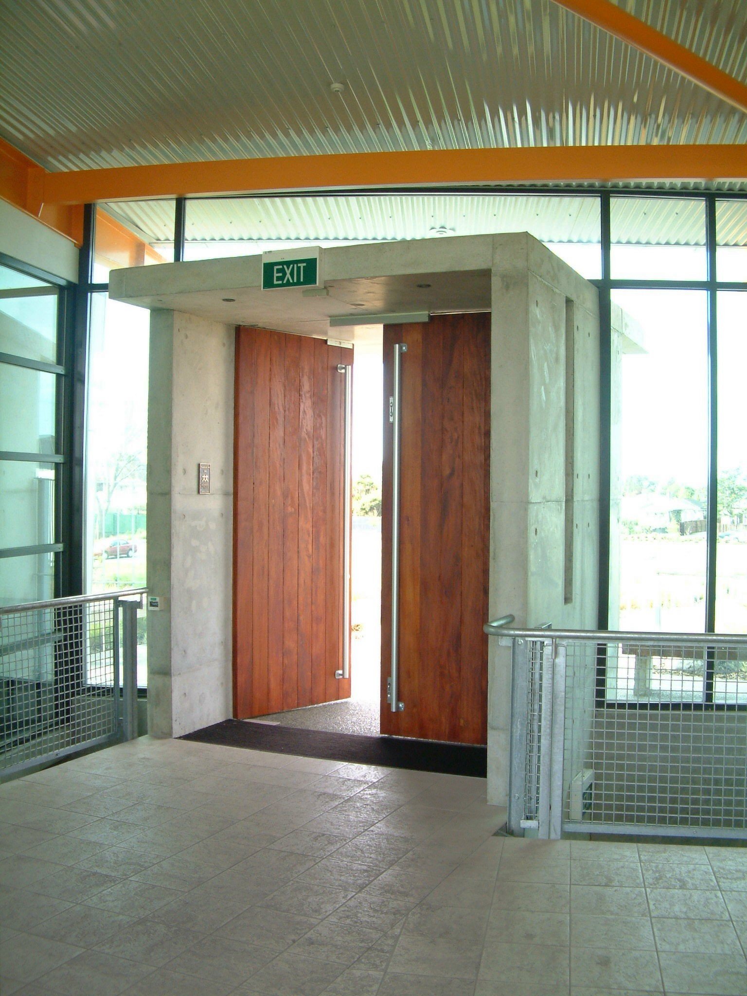 Interior view of a building entrance with open wooden doors, concrete and glass walls, and an illuminated green 'EXIT' sign