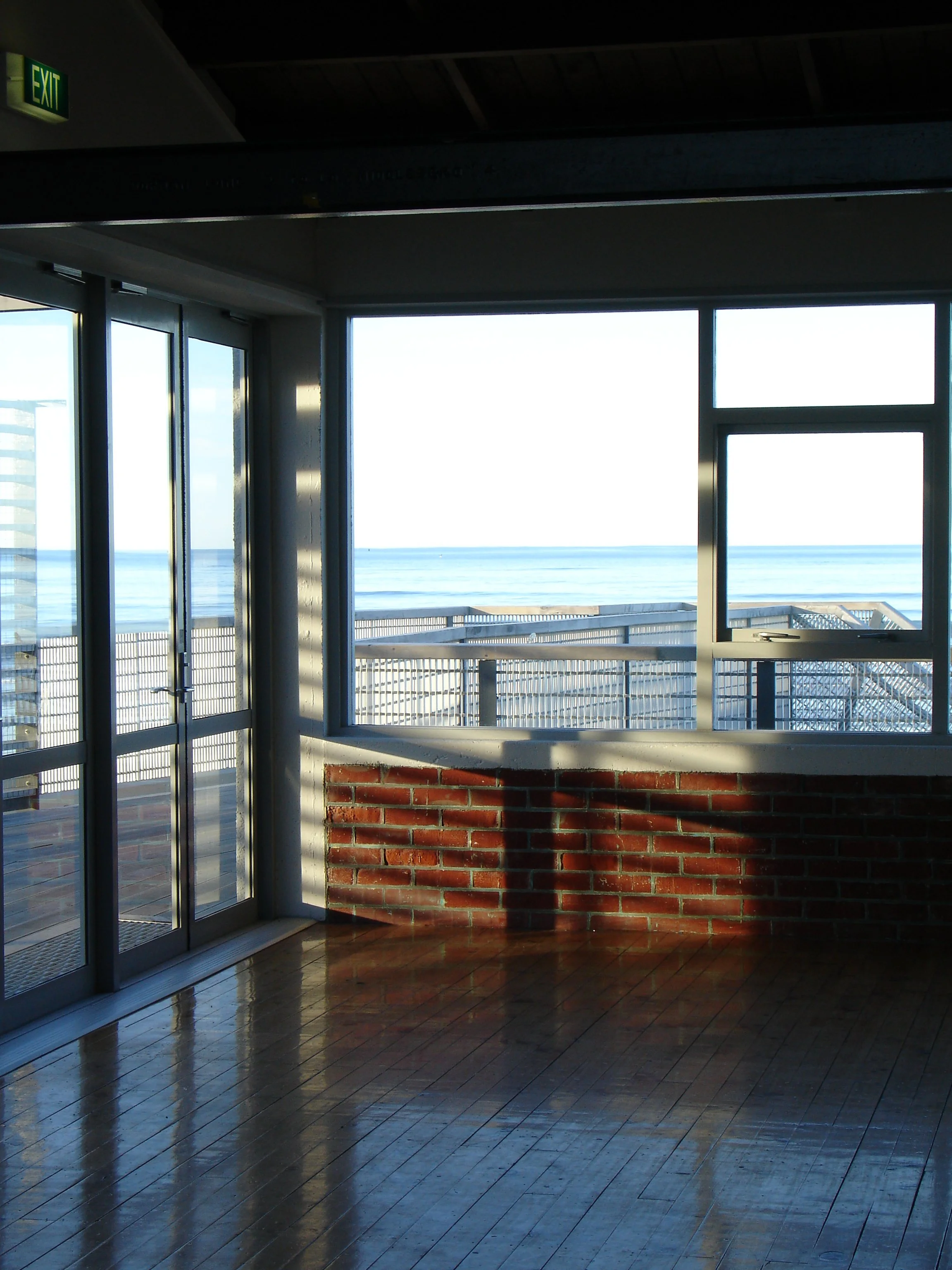Interior of a room with large windows and a glass door, overlooking a deck and the ocean. Sunlight casts shadows on the wooden floor and brick wall.