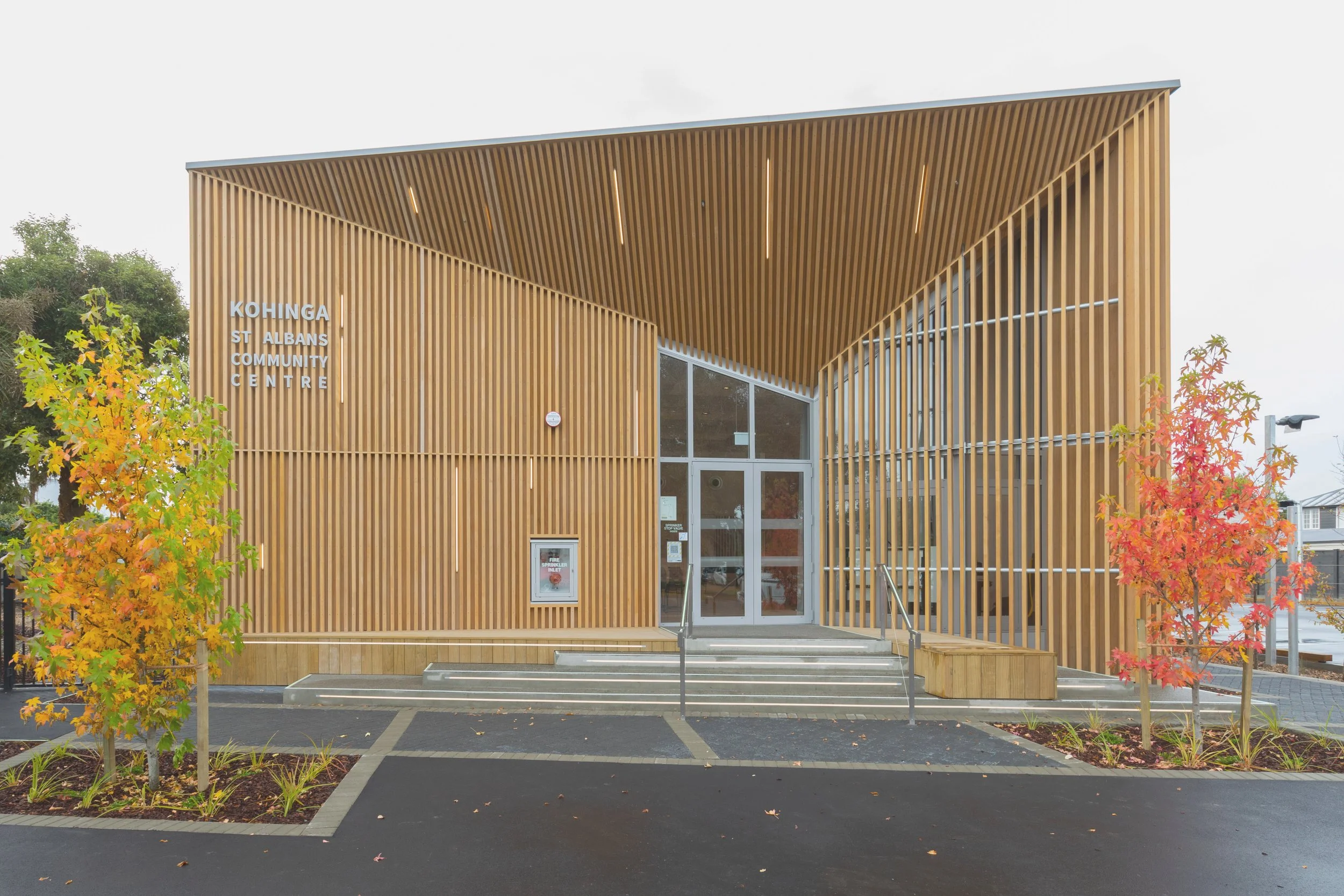 Front view of Kohinga St Albans Community Centre building with wooden slat facade, glass door entrance, steps, and surrounding trees with autumn leaves.
