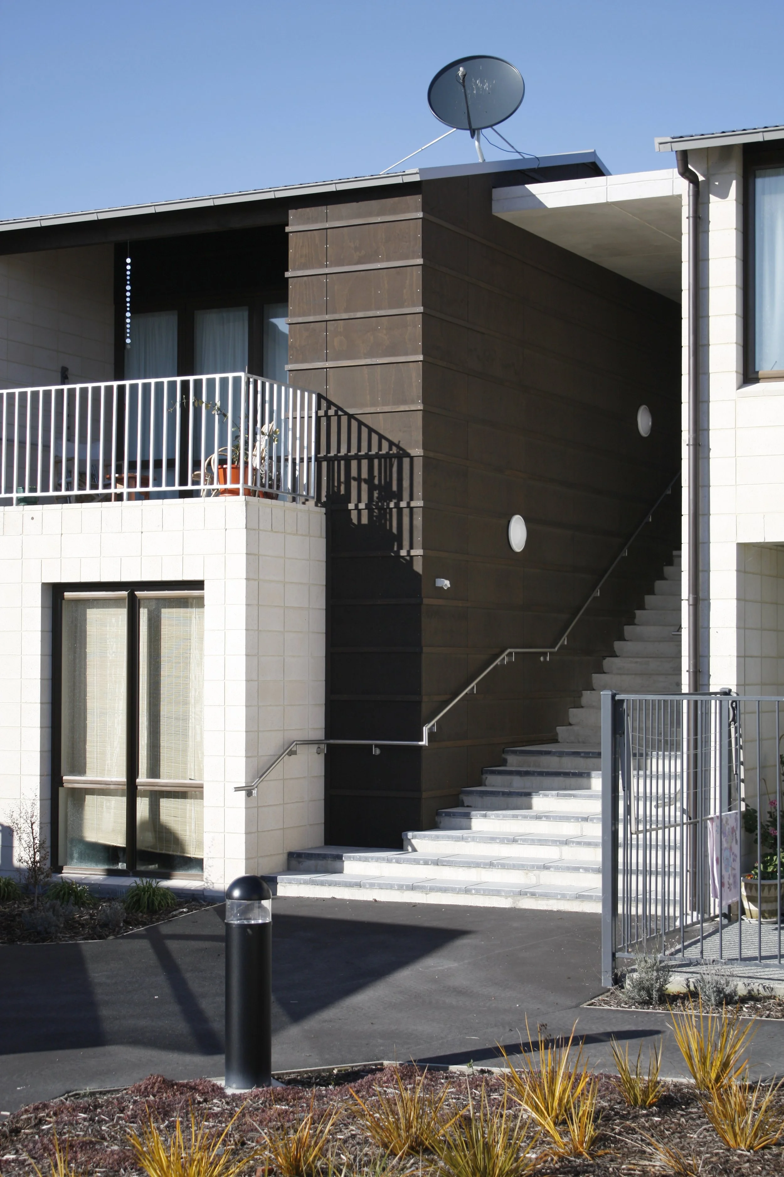 Exterior view of a modern building with stairs, a balcony with plants, satellite dish on the roof, and gated entrance on a sunny day.