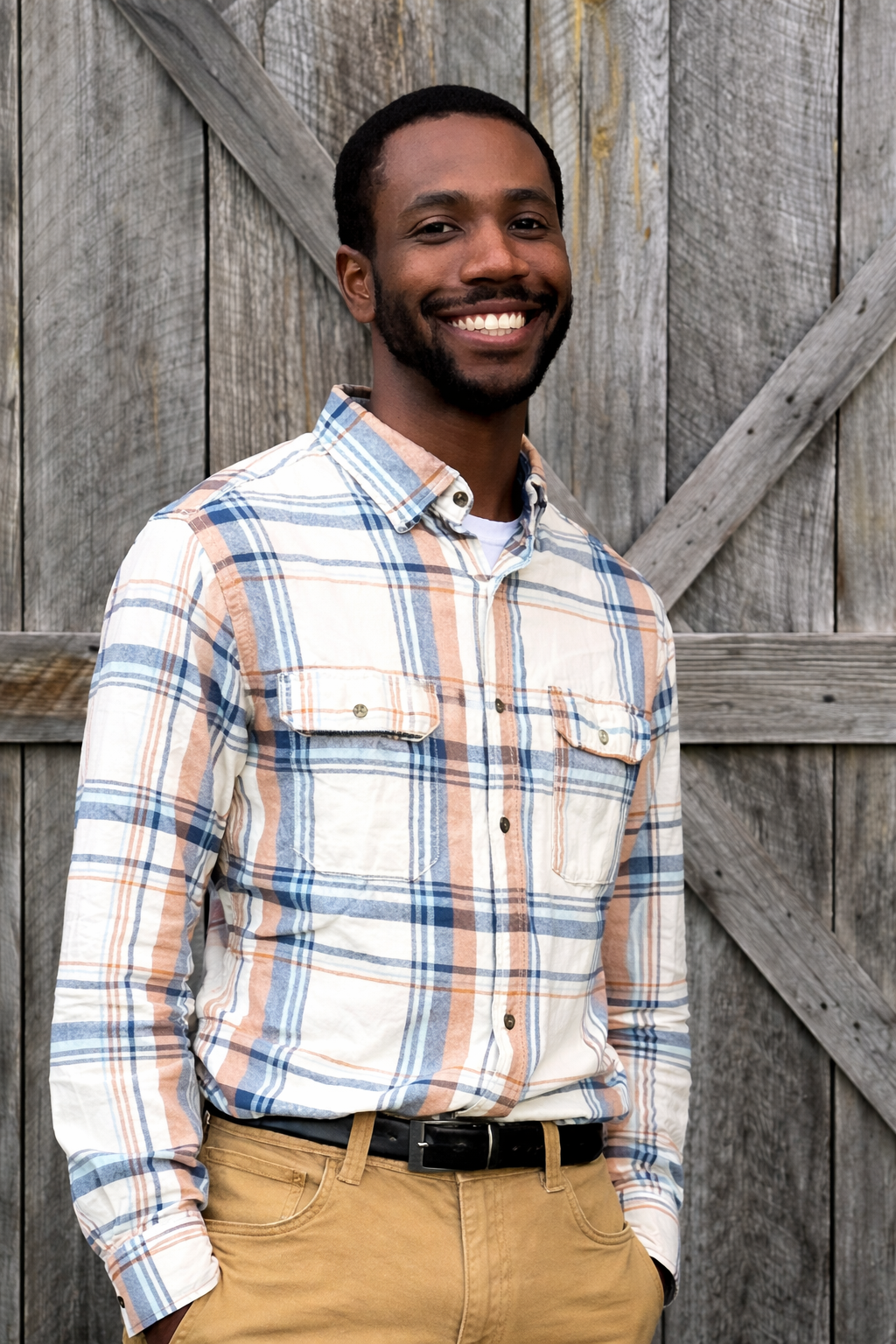 A man with a short beard and mustache wearing a plaid shirt and khaki pants, smiling while standing in front of a wooden barn door.