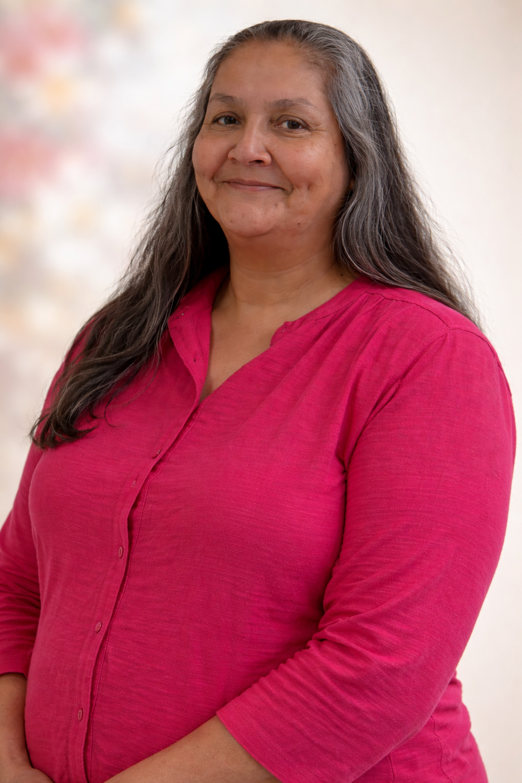 Portrait of a woman with long gray hair, smiling, wearing a bright pink shirt, standing against a blurred light-colored background.