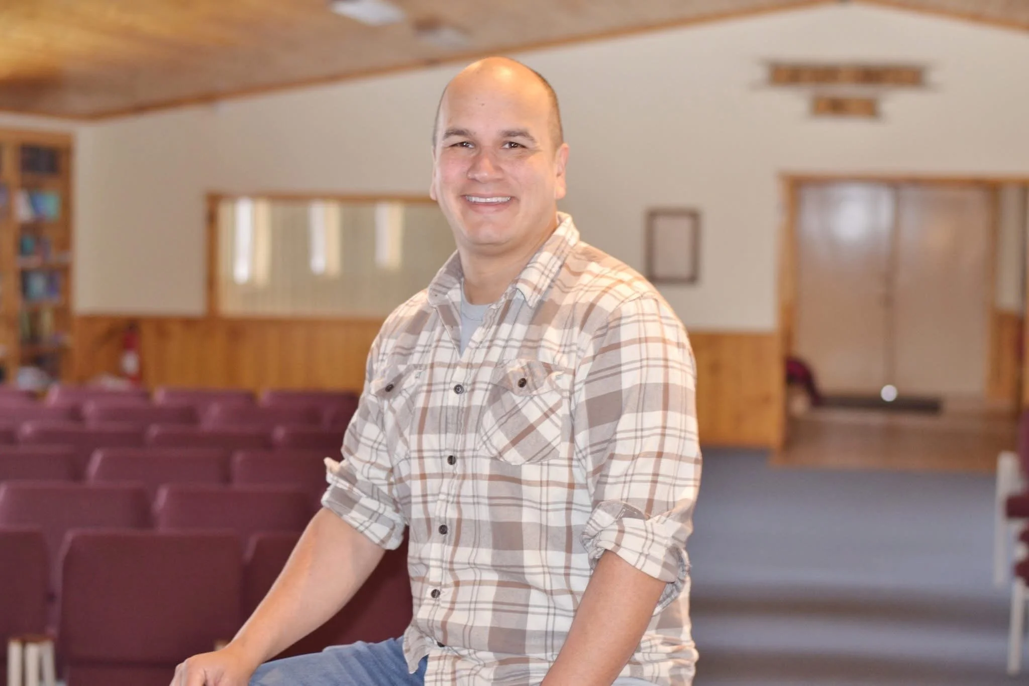 A smiling man in a plaid shirt sitting in an empty auditorium with rows of maroon chairs and wooden walls.