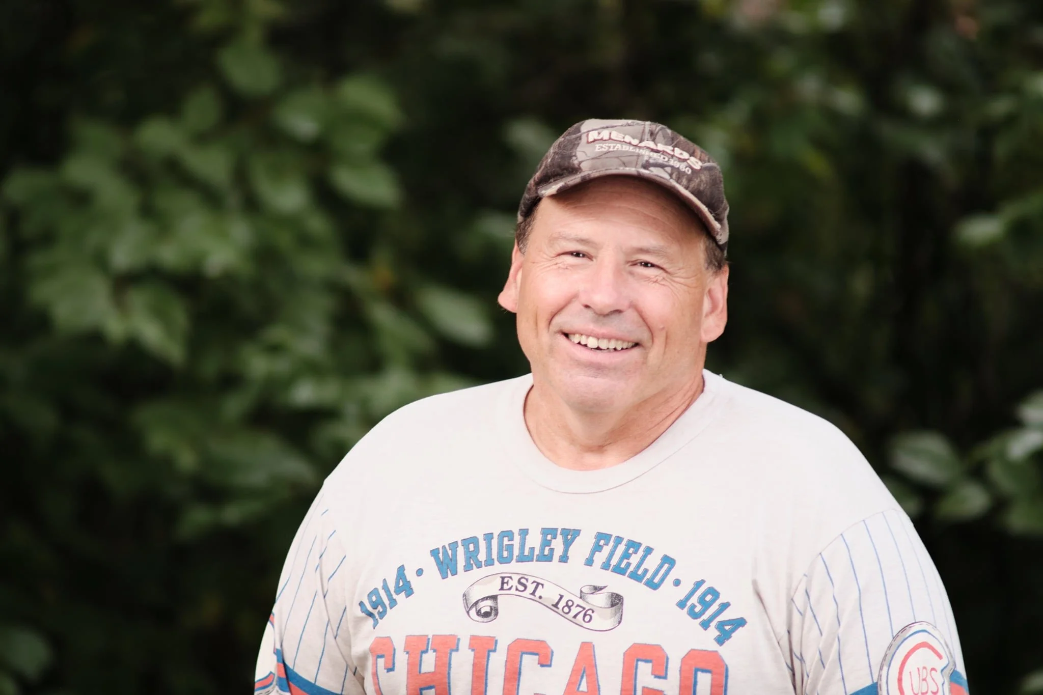 A man smiling outdoors with a background of green foliage, wearing a white T-shirt and a camouflage baseball cap.