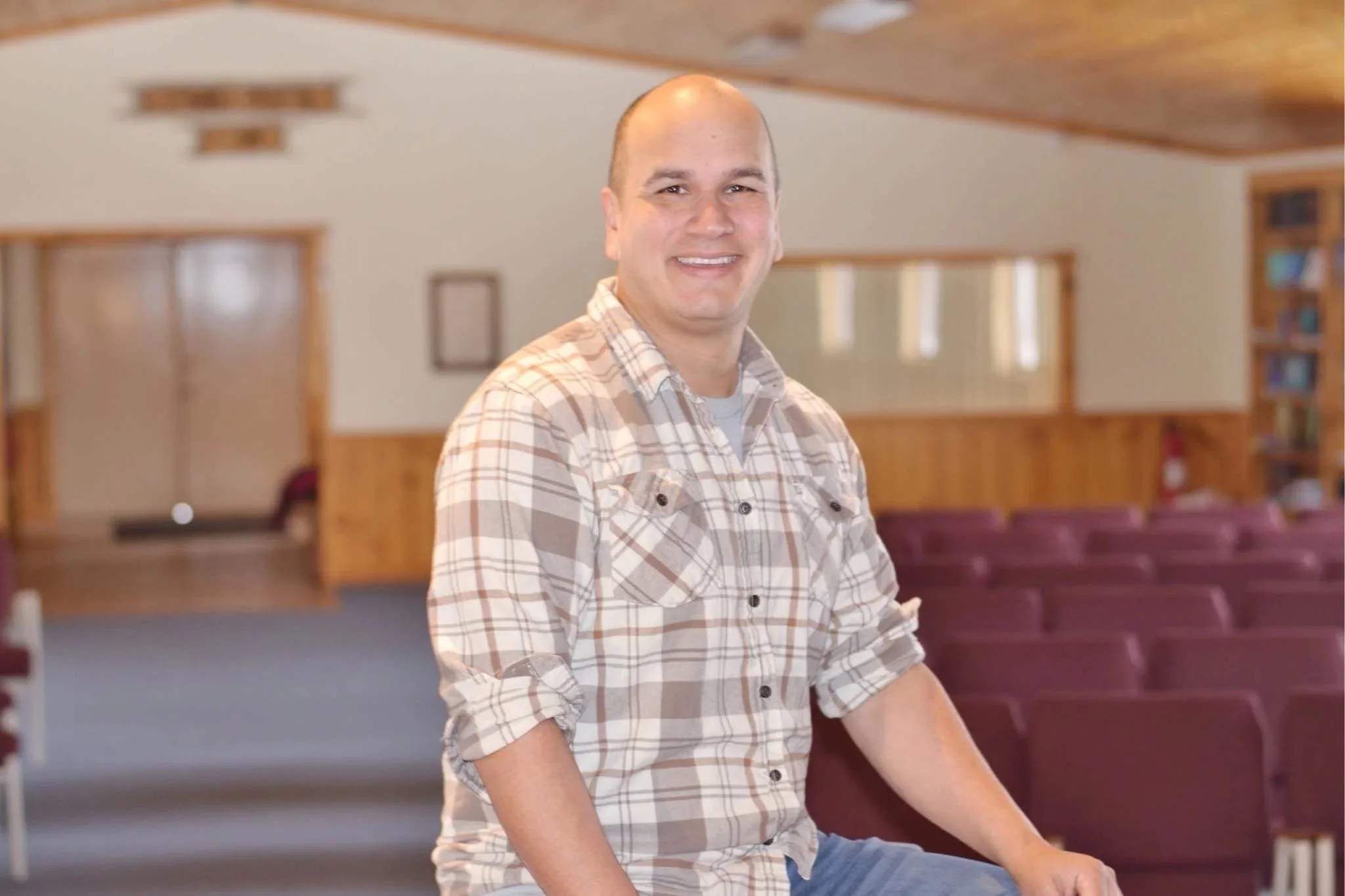 A smiling man in a plaid shirt sitting in an empty church or auditorium with rows of purple chairs and wooden paneling.