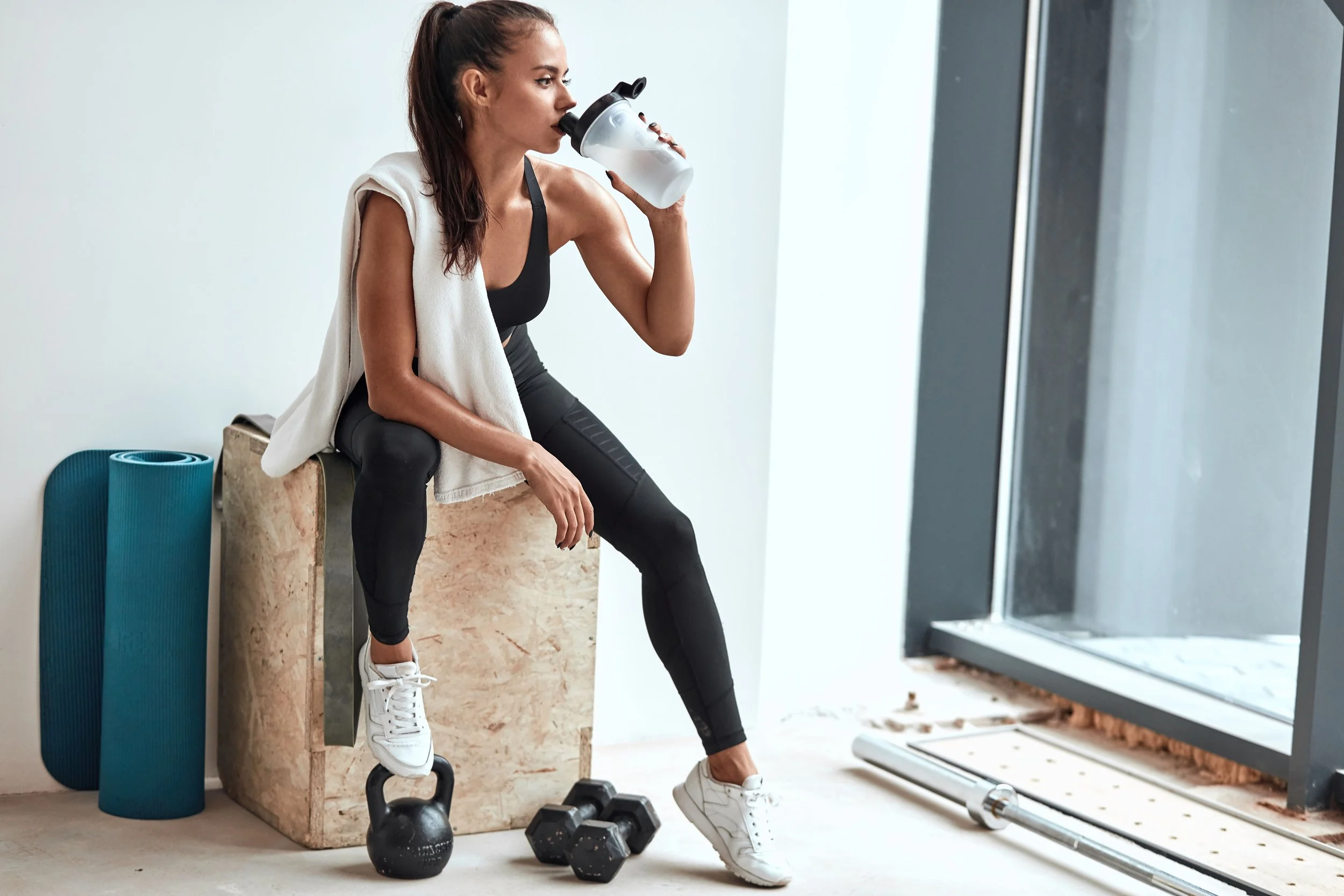 Une femme faisant une pause après une séance d'entraînement, buvant de l'eau et assise sur un banc avec des haltères et un tapis de yoga à côté, dans une salle de sport moderne.