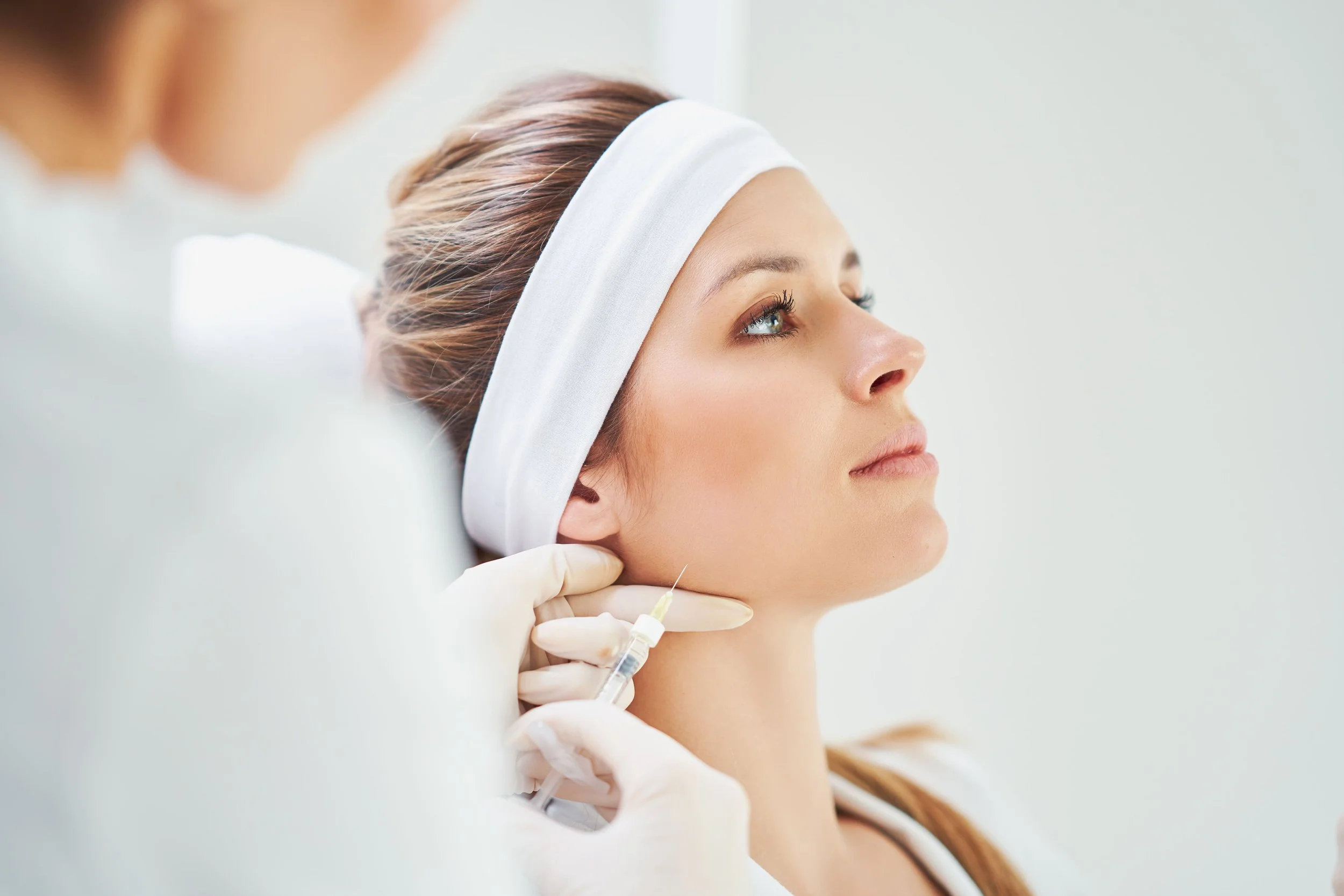 Femme recevant une injection lors d'une procédure cosmétique ou médicale, portant un bandeau blanc, avec une praticienne portant des gants blancs.
