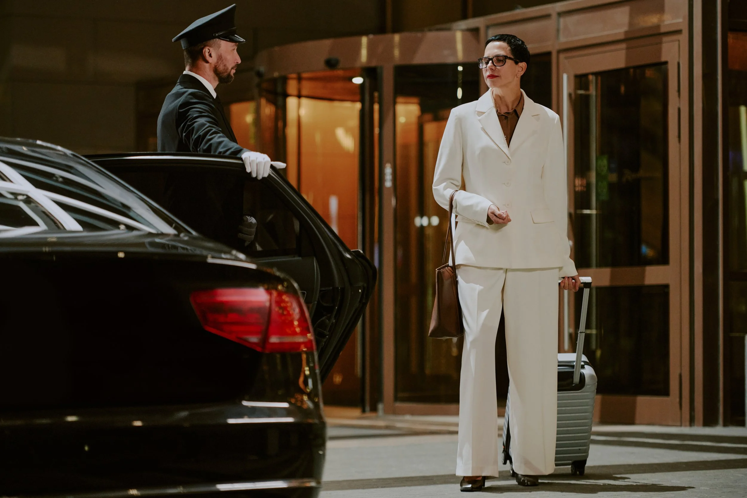 Une femme en tailleur blanc avec des lunettes, tenant une valise, parlant à un porteur en uniforme qui ouvre la porte d'une voiture noir devant un bâtiment.