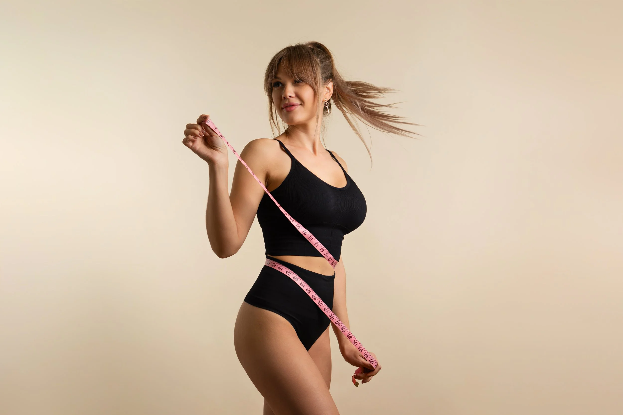 Une femme souriante en vêtements de sport noirs, mesurant sa taille avec un ruban à mesurer rose, devant un fond beige clair.