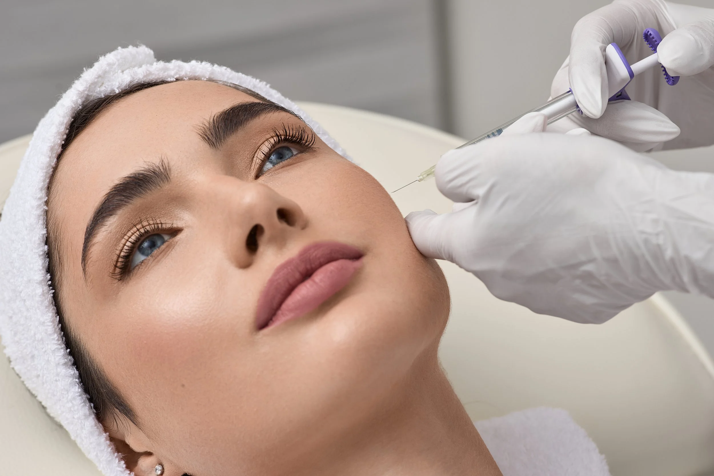 Femme recevant une injection esthétique au visage, cloche de coton sur la tête, main en gants blancs tenant une seringue.