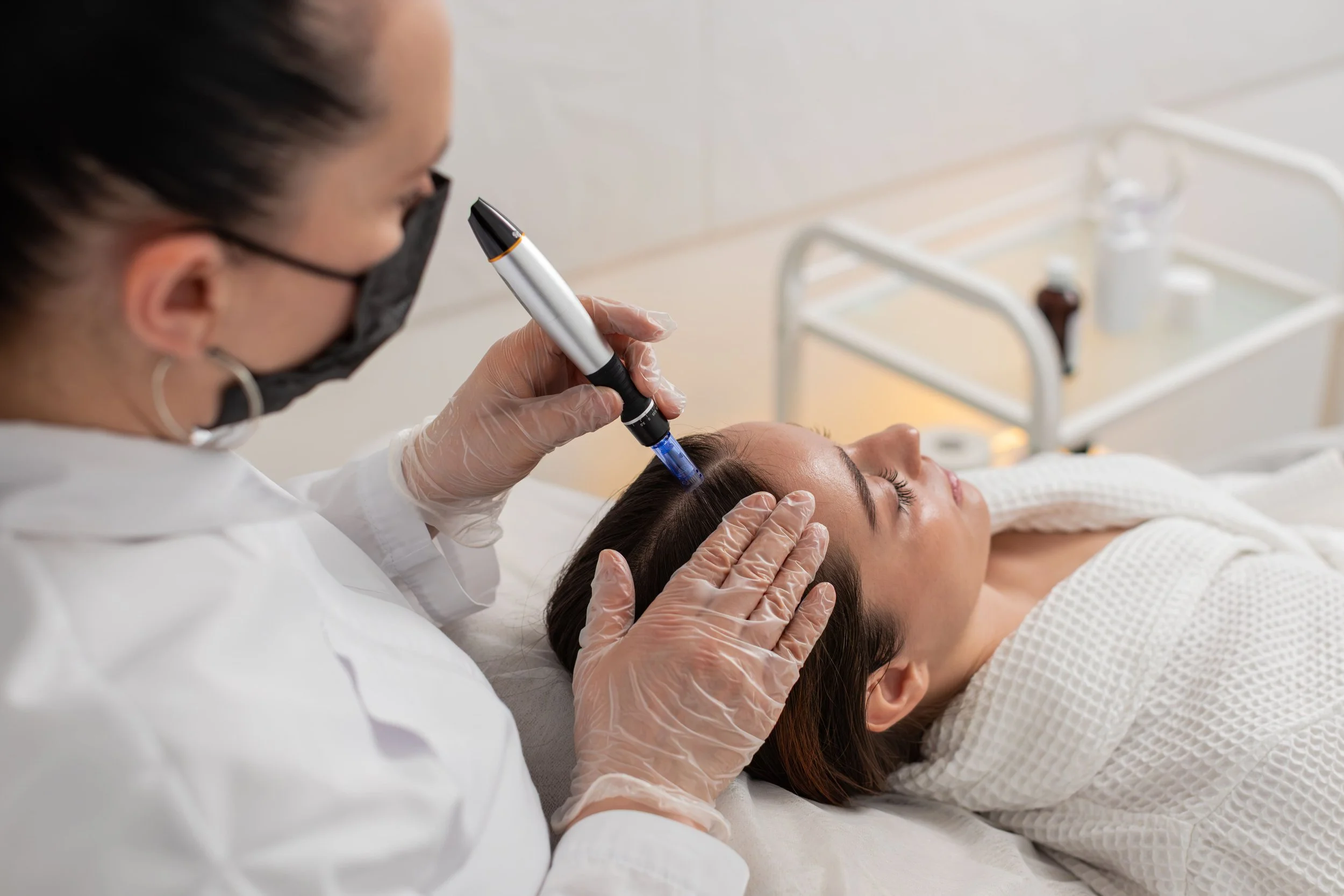 Une esthéticienne ou une médecin utilise un appareil pour une procédure sur le visage d'une femme allongée sur une table d'examen, dans un environnement médical ou de soins de beauté.