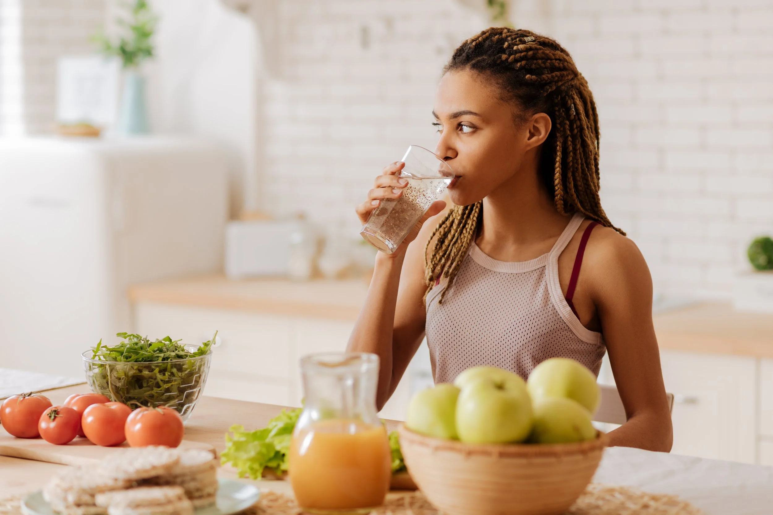 Une femme aux cheveux en tresses brunes boit un verre d'eau dans une cuisine lumineuse, entourée de fruits et légumes comme des tomates, des pommes, un saladier de salade et un jus d'orange sur la table.