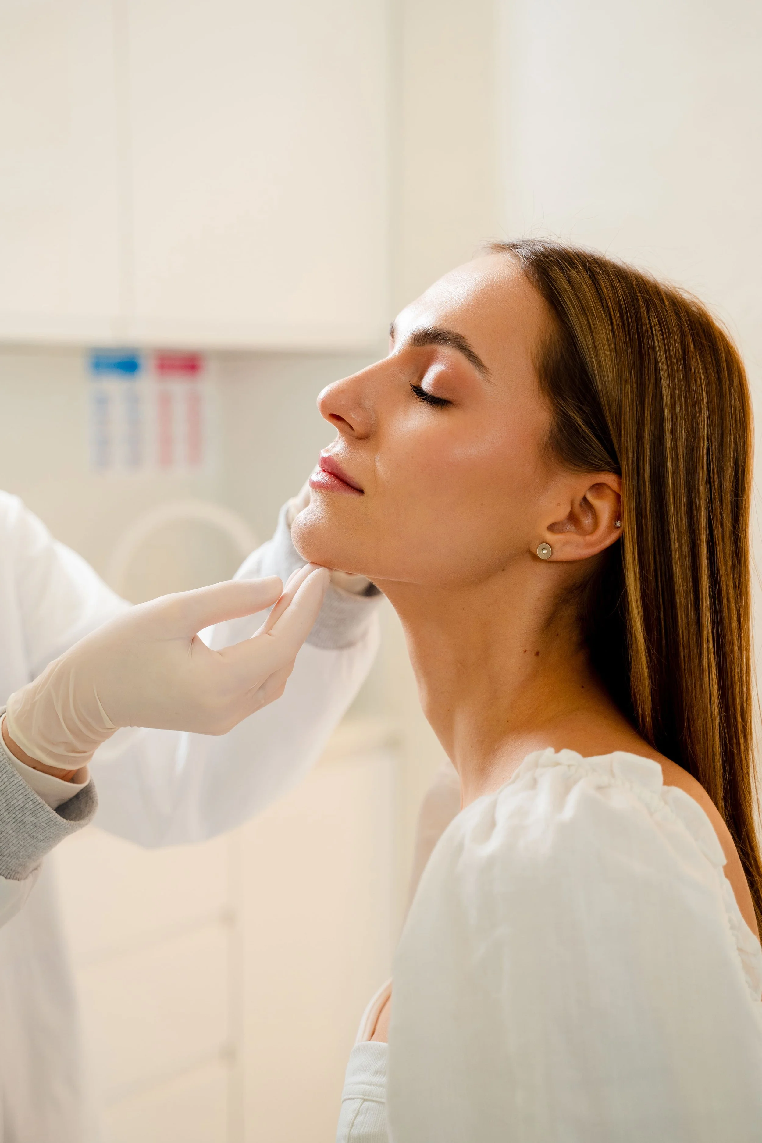Femme ayant les yeux fermés, recevant une injection ou un traitement médical dans le cou, opérée par un professionnel de santé portant des gants, dans un environnement clinique.
