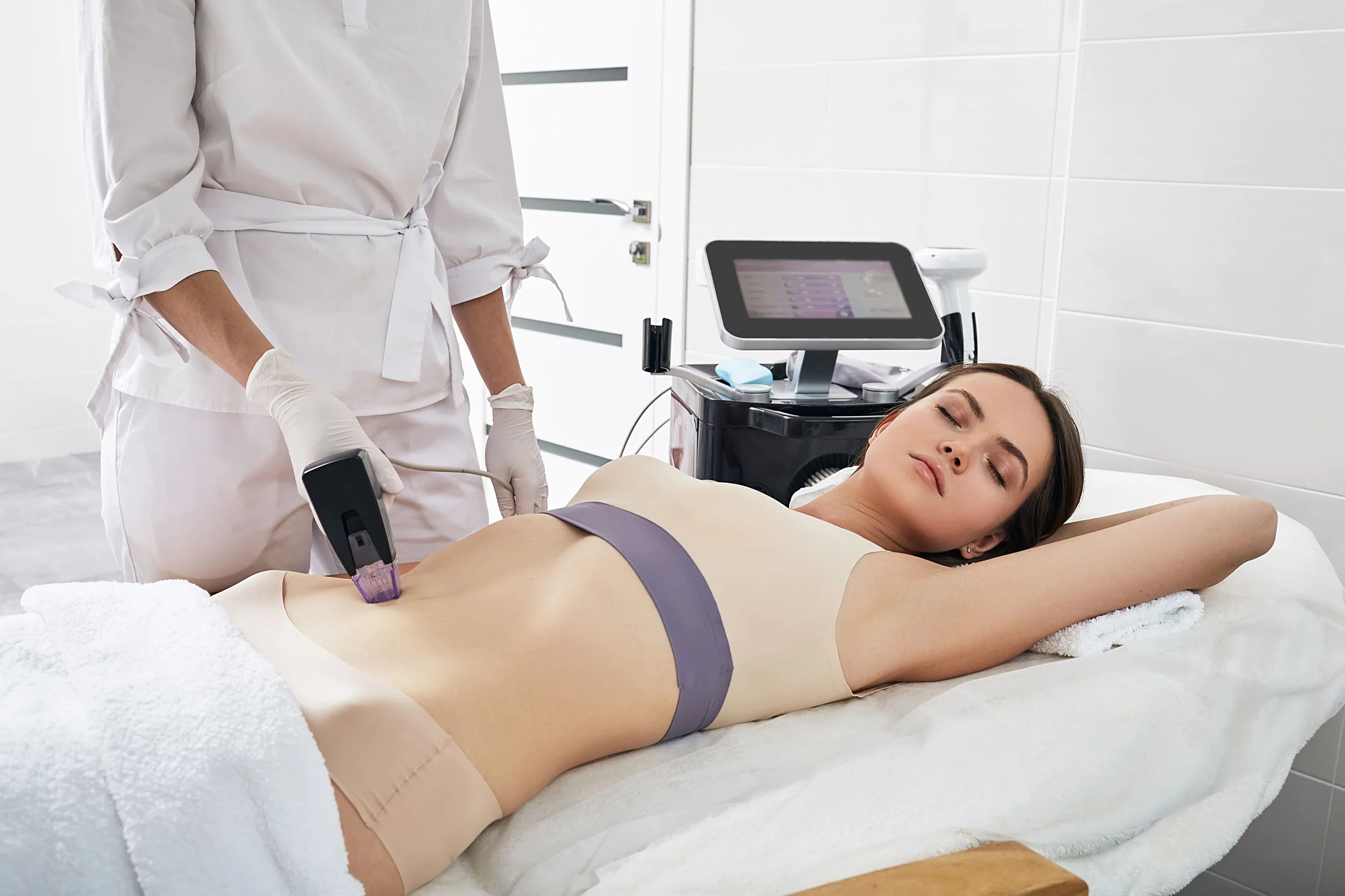 Une femme allongée sur une table de soin recevant un traitement avec un appareil électronique porté à son abdomen, une professionnelle portant des gants blancs manipule l'appareil dans une pièce blanche.