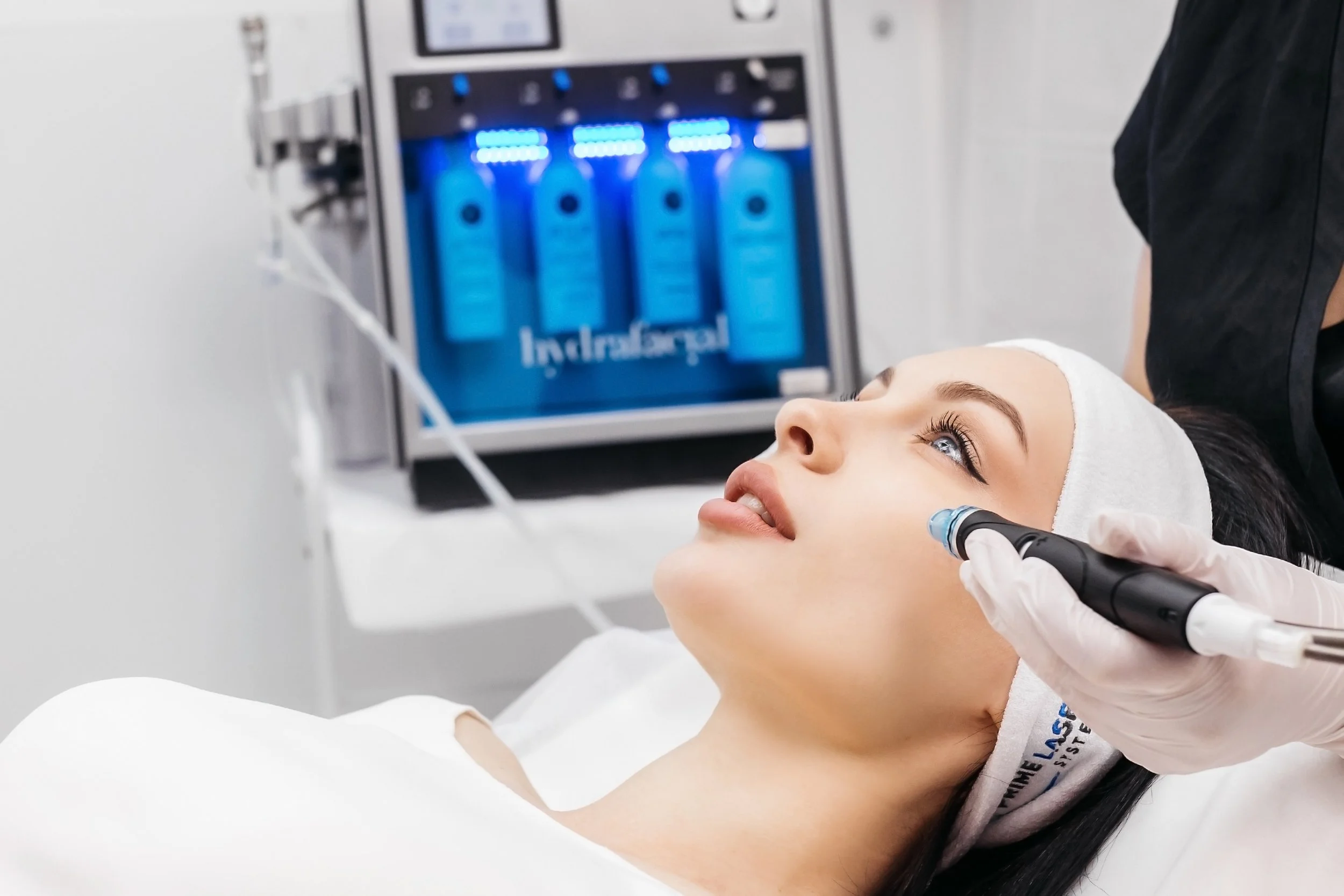 Une femme allongée recevant un traitement médical au visage avec un appareil noir et un professionnel portant un gant blanc. En arrière-plan, une machine avec des bouteilles d'eau bleues éclairées en bleu.
