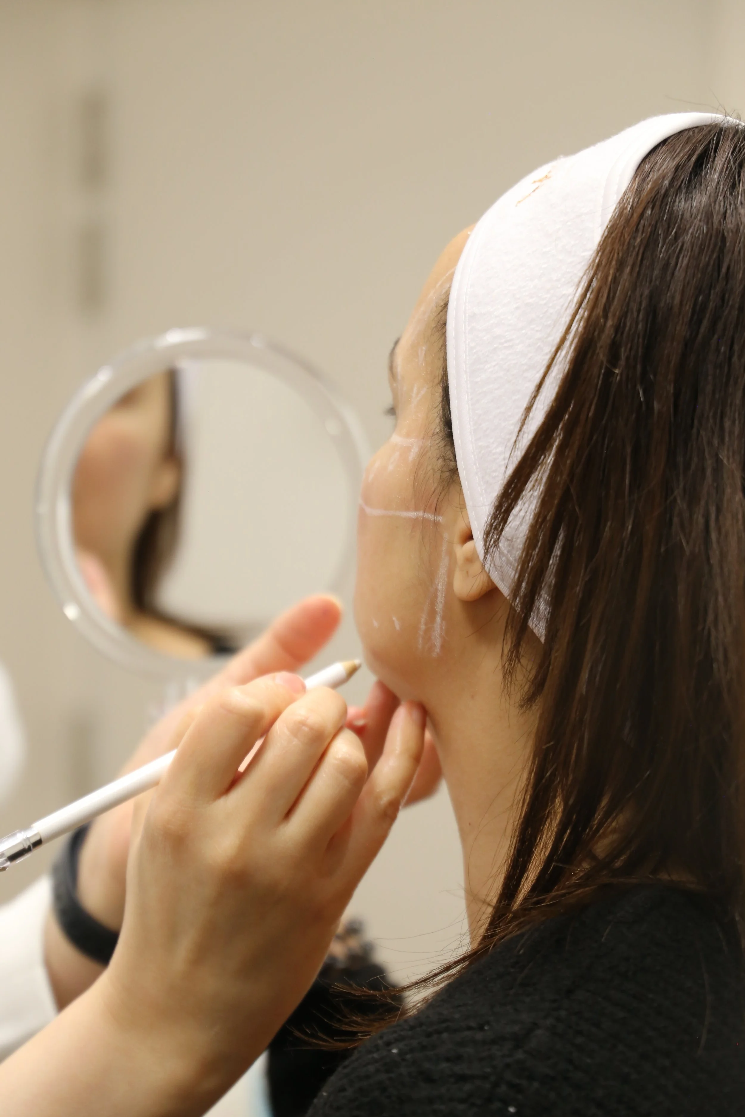 Une femme se fait maquiller autour de la zone des yeux avec un pinceau. Elle porte un bandeau blanc et a les cheveux bruns, longs, et lisses. La scène se déroule probablement dans un salon de beauté ou un institut de maquillage.