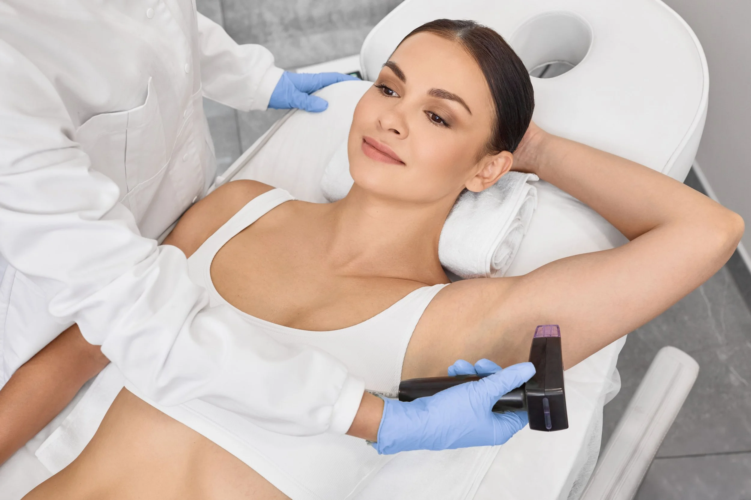 Une femme allongée sur une table d'examen, une professionnelle de santé en blouse blanche et en gants bleus lui administre une injection dans le bras.